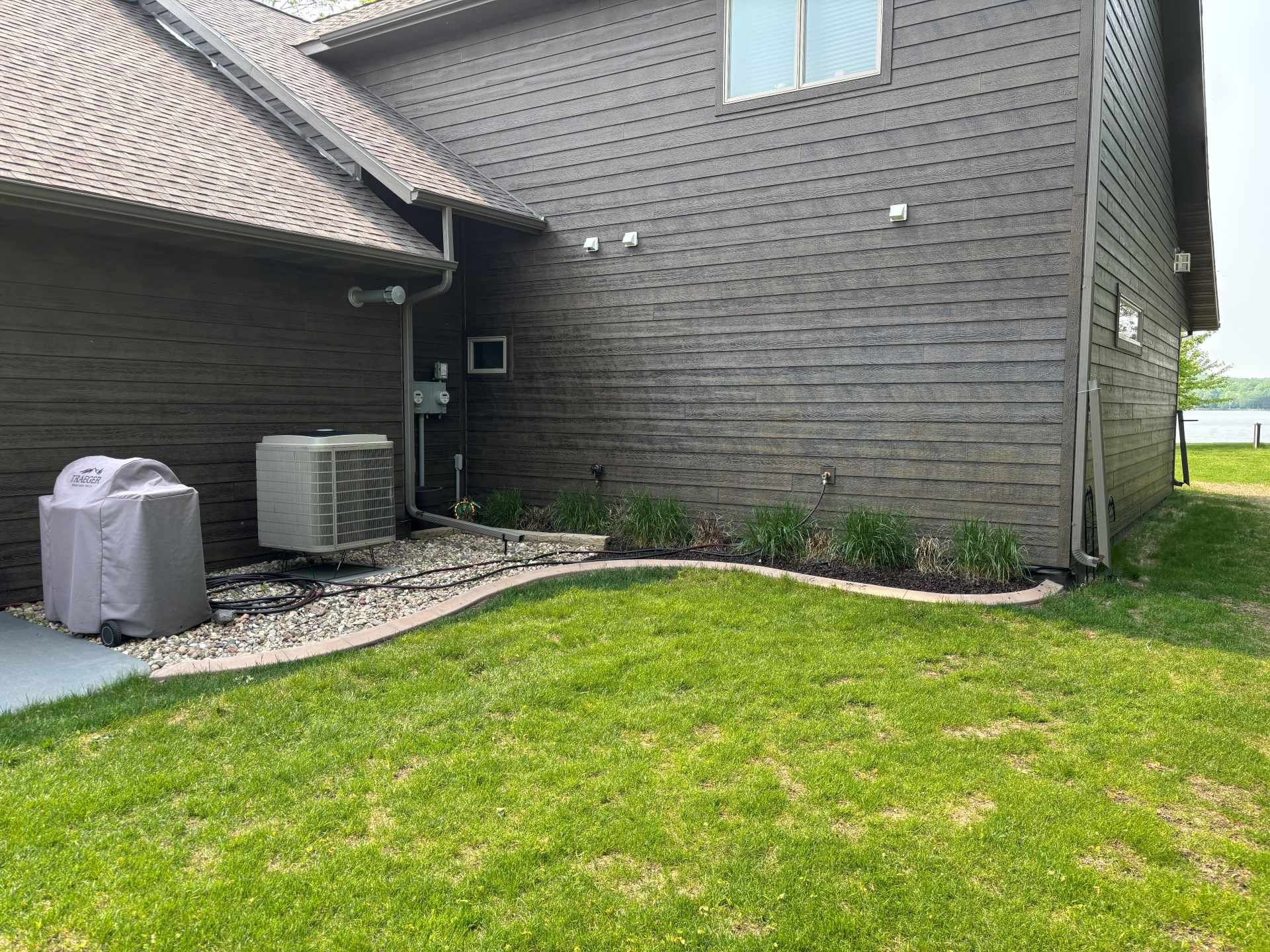 A dark-sided house exterior with a green lawn, rock landscaping, a covered grill, and an air conditioning unit.