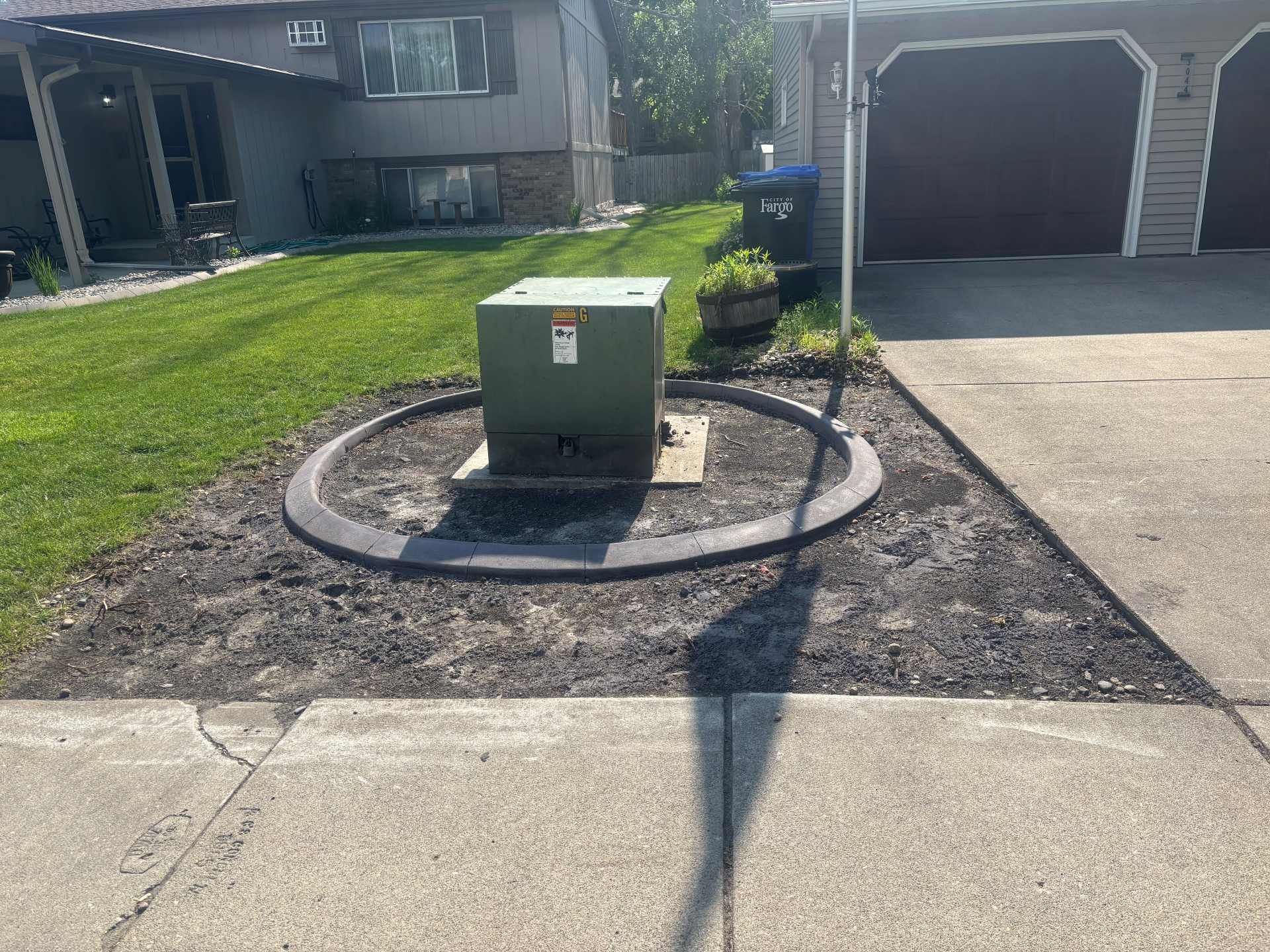 A green utility transformer box sits in a landscaped circle of mulch, surrounded by a gray driveway and a grassy yard.