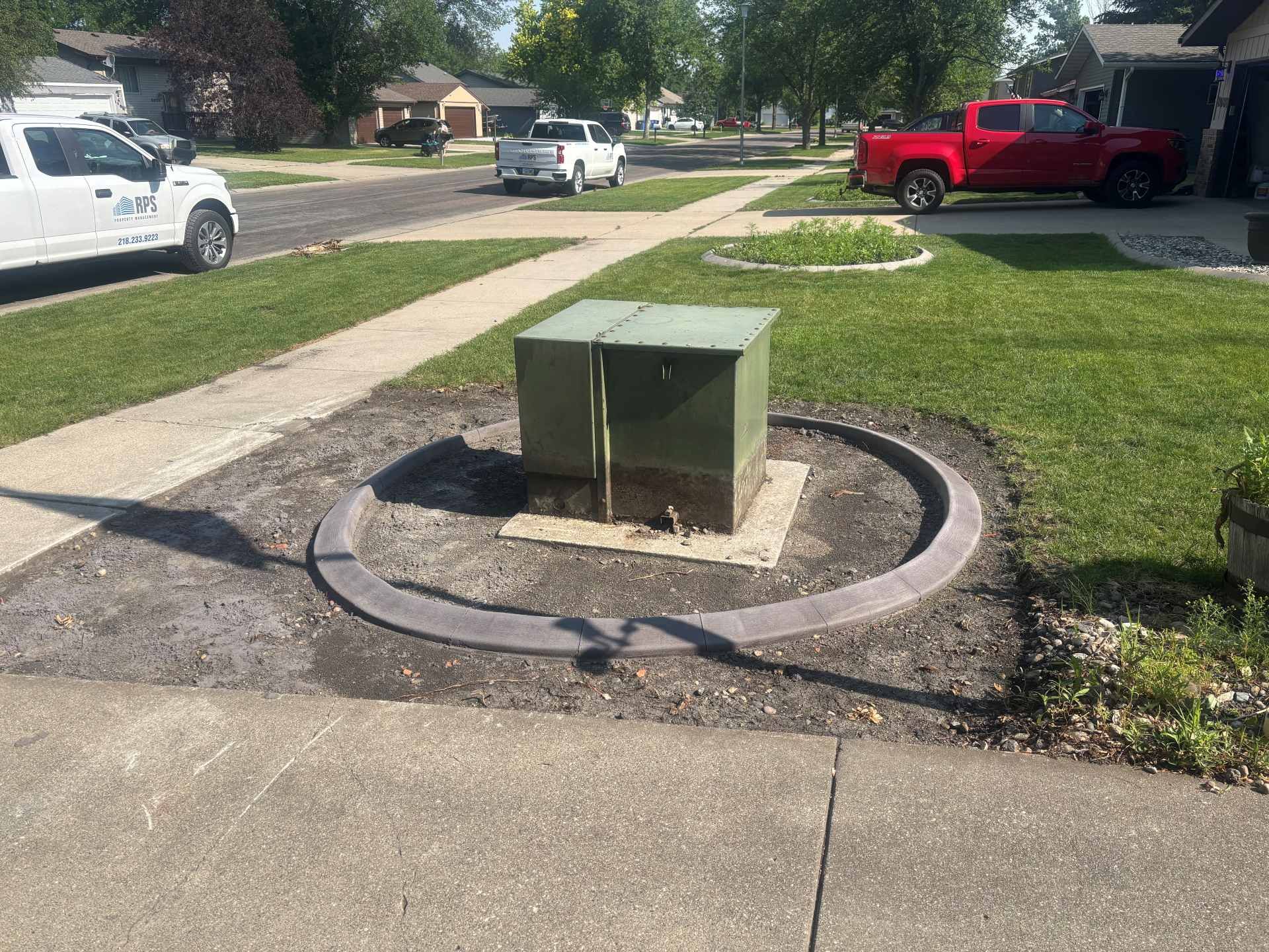 A green utility box sits in a circular concrete border on a suburban lawn, flanked by a sidewalk and parked vehicles.