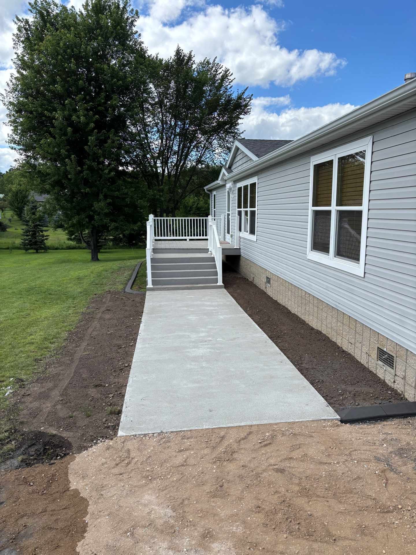 A concrete sidewalk leads to a raised wooden deck with white railings attached to a light gray, single-story house.