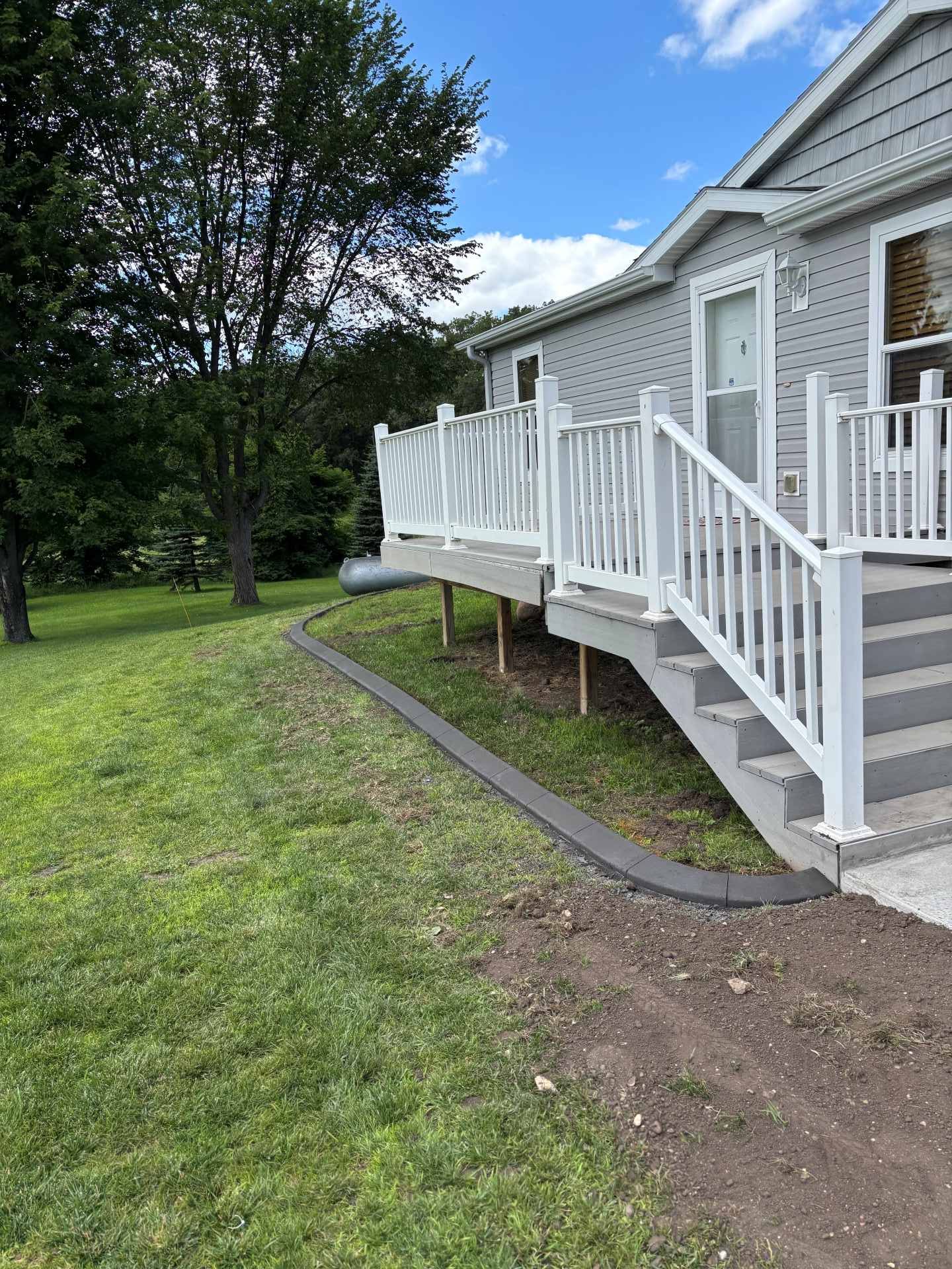 A grey house with a white railing deck and a newly installed curved concrete landscape border edging the lawn.