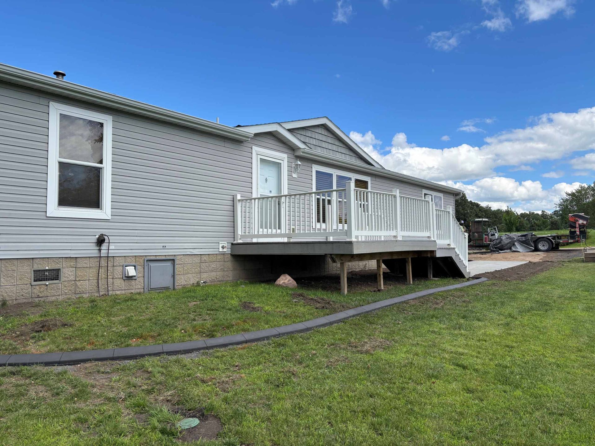 A gray manufactured home with a white deck, set against a sunny, blue sky in a grassy yard.