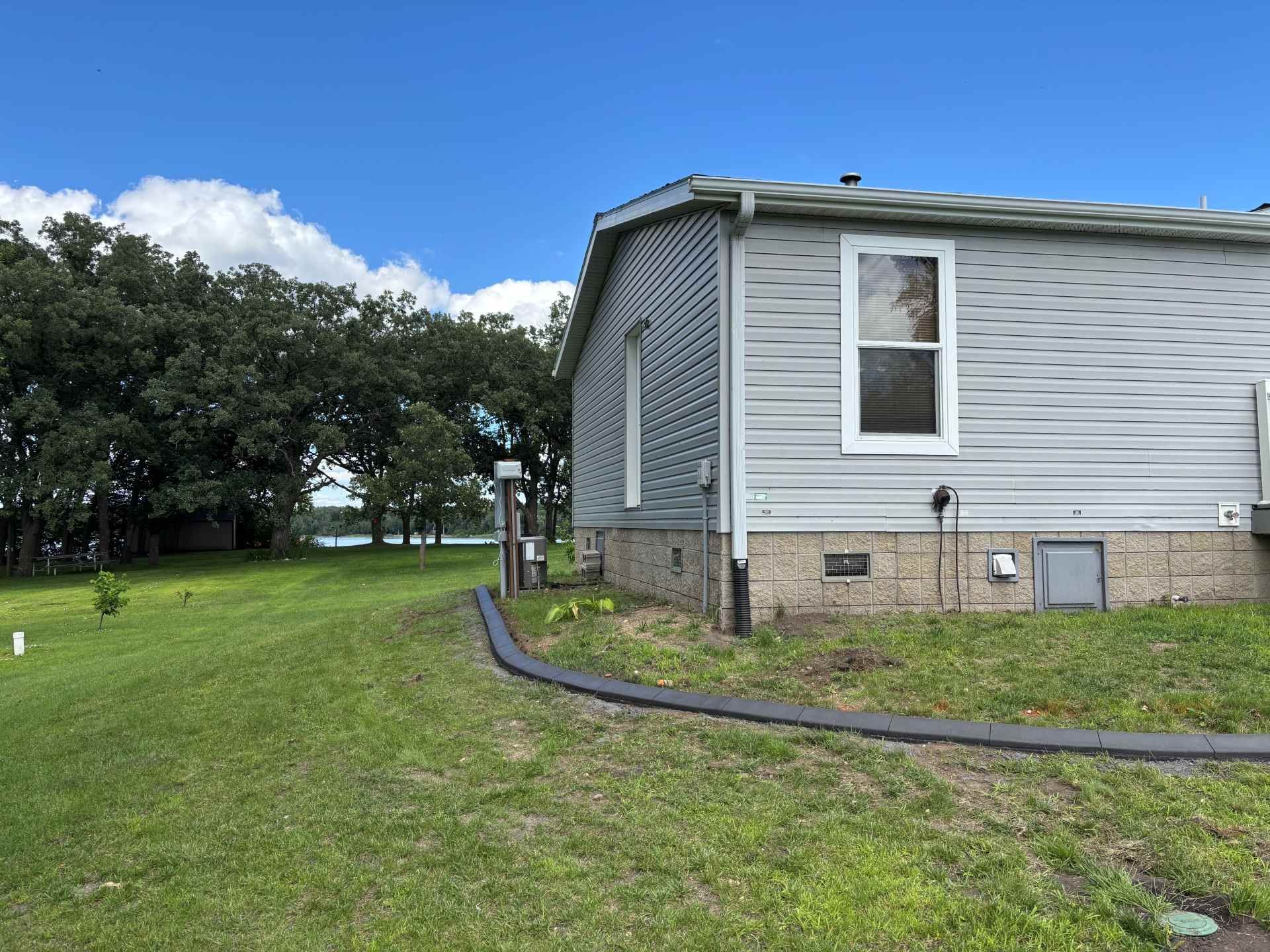 A grey house with a concrete foundation sits on a grassy lawn near trees and a lake under a clear blue sky.