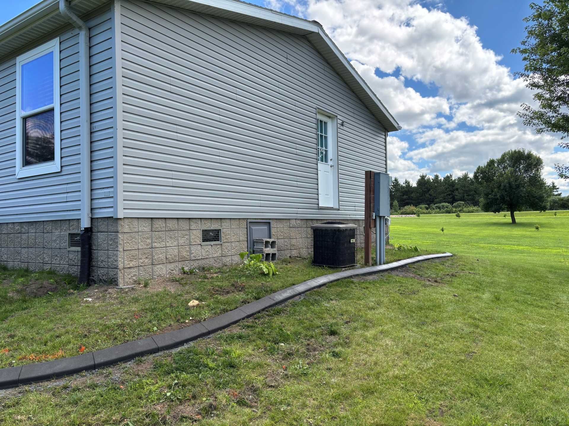 A light gray home with horizontal siding sits on a concrete block foundation in a grassy yard under a blue sky.