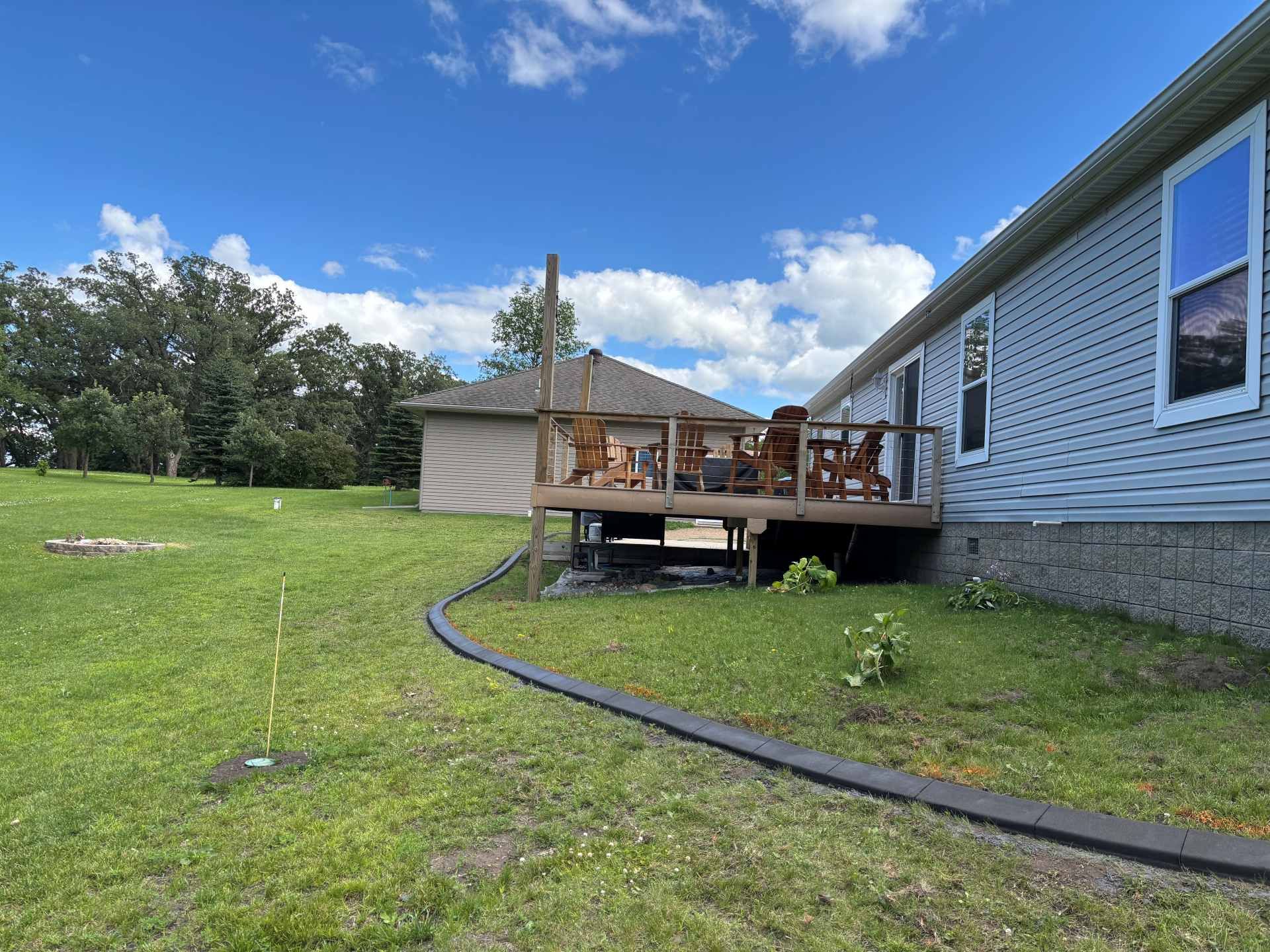 A wooden deck attached to a gray house, bordered by a black landscape edging on a green lawn under a sunny, blue sky.