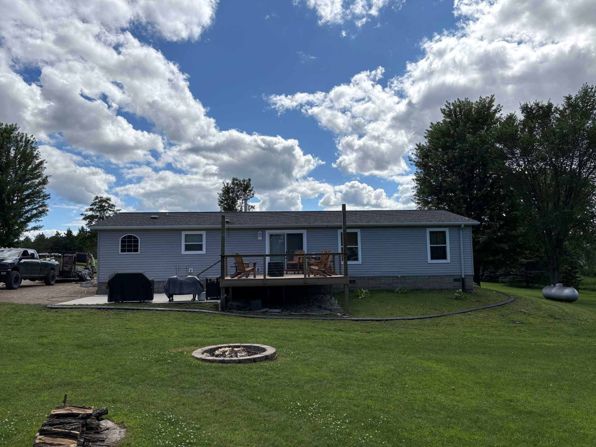 A light blue manufactured home with a wooden deck, a fire pit, and a grassy yard under a partly cloudy sky.
