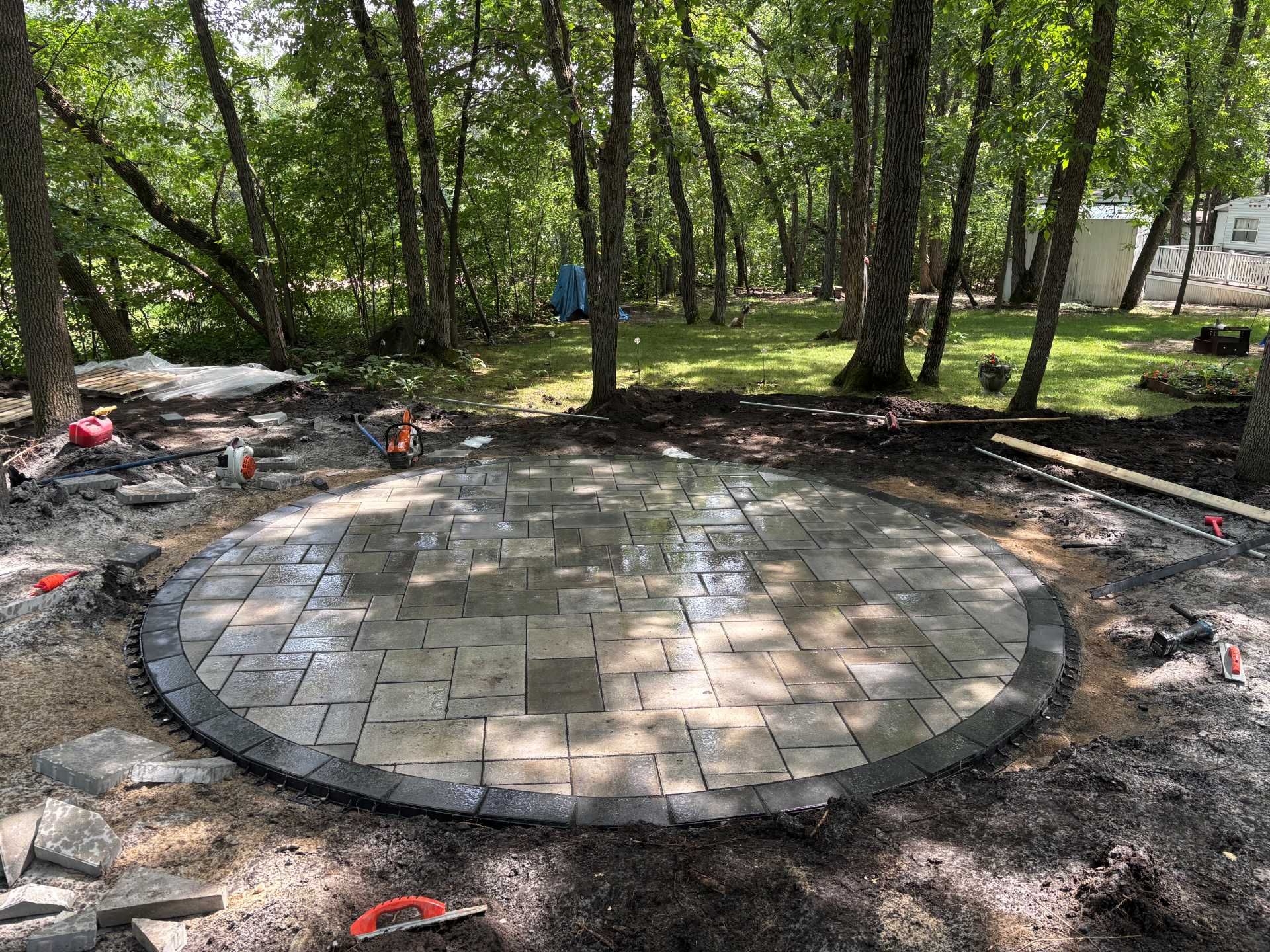 A circular stone patio under construction in a wooded backyard, featuring a dark border and light-colored paver center.