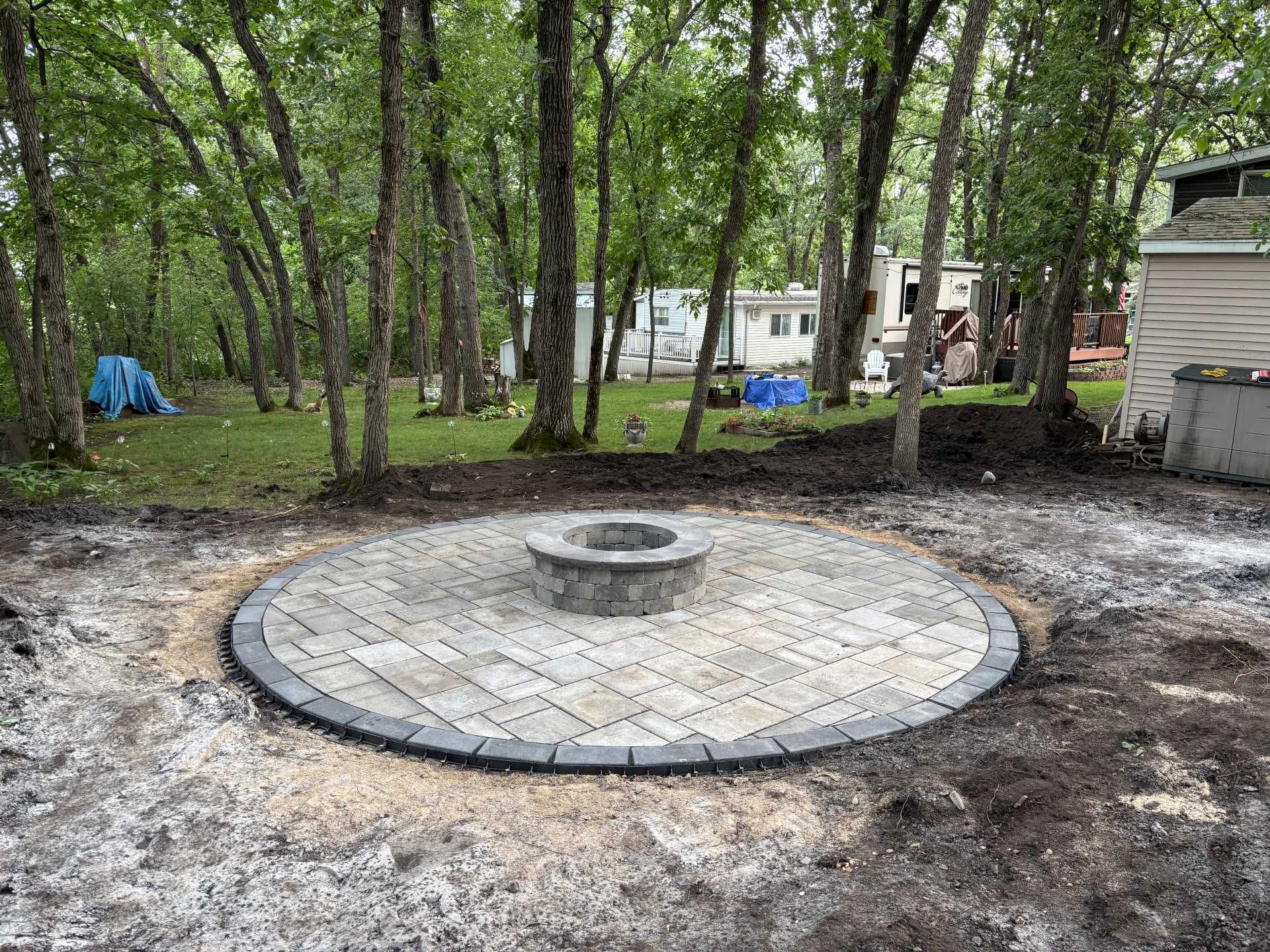 A circular stone patio with a central fire pit sits in a grassy, wooded yard near residential trailers.