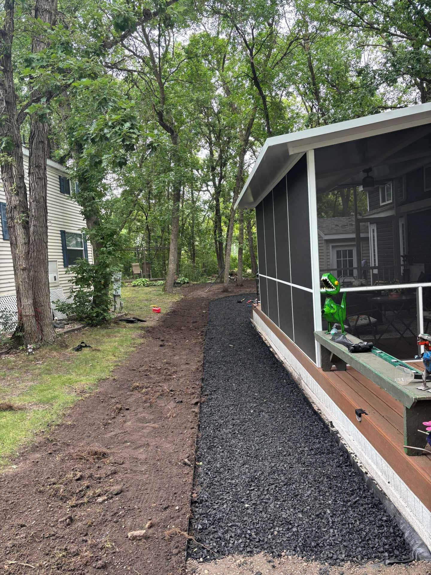 A gravel pathway runs along the side of a screened-in porch next to an area of tilled dirt in a wooded backyard.