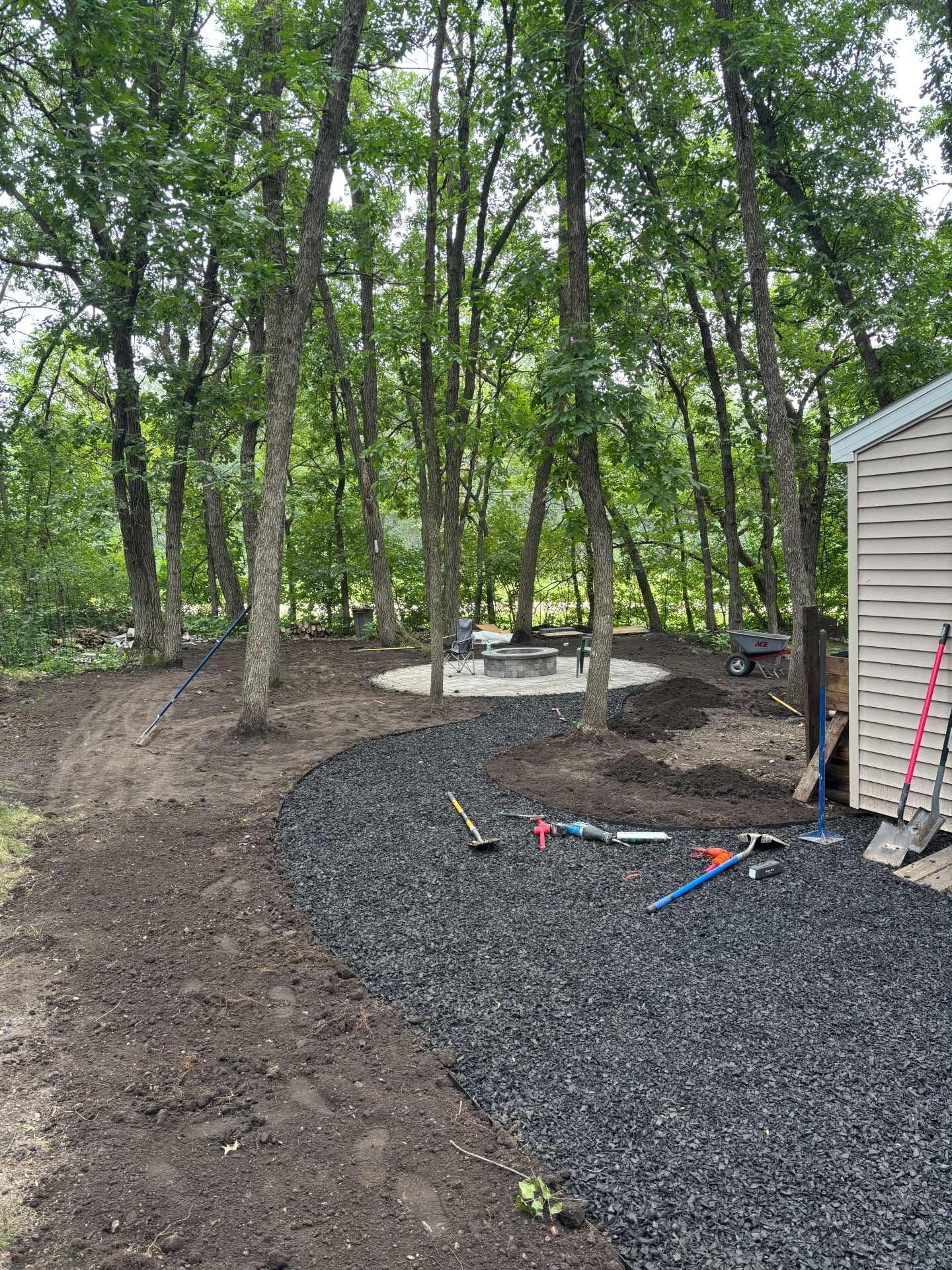 A backyard landscape project showing a freshly laid gravel path winding toward a stone fire pit area among trees.