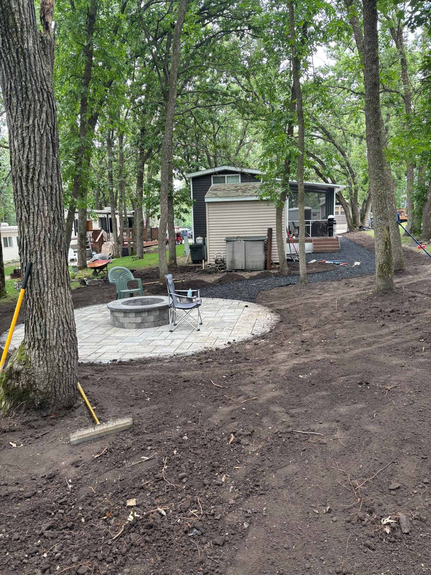 A circular stone fire pit sits in a cleared, wooded yard in front of a small cabin, with a rake leaning against a tree.