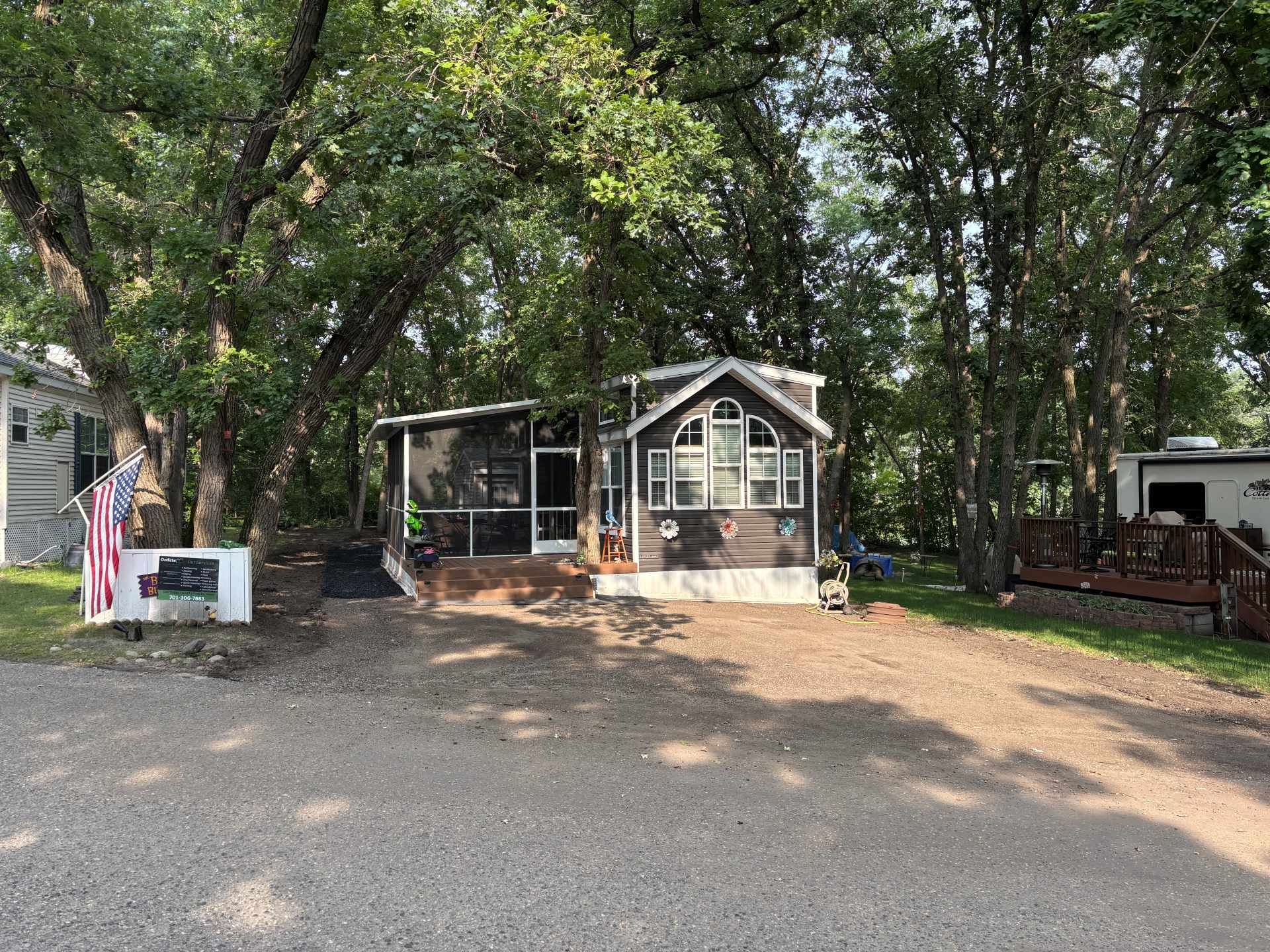 A dark-sided cabin with a screened porch sits in a wooded area with a gravel driveway and an American flag.