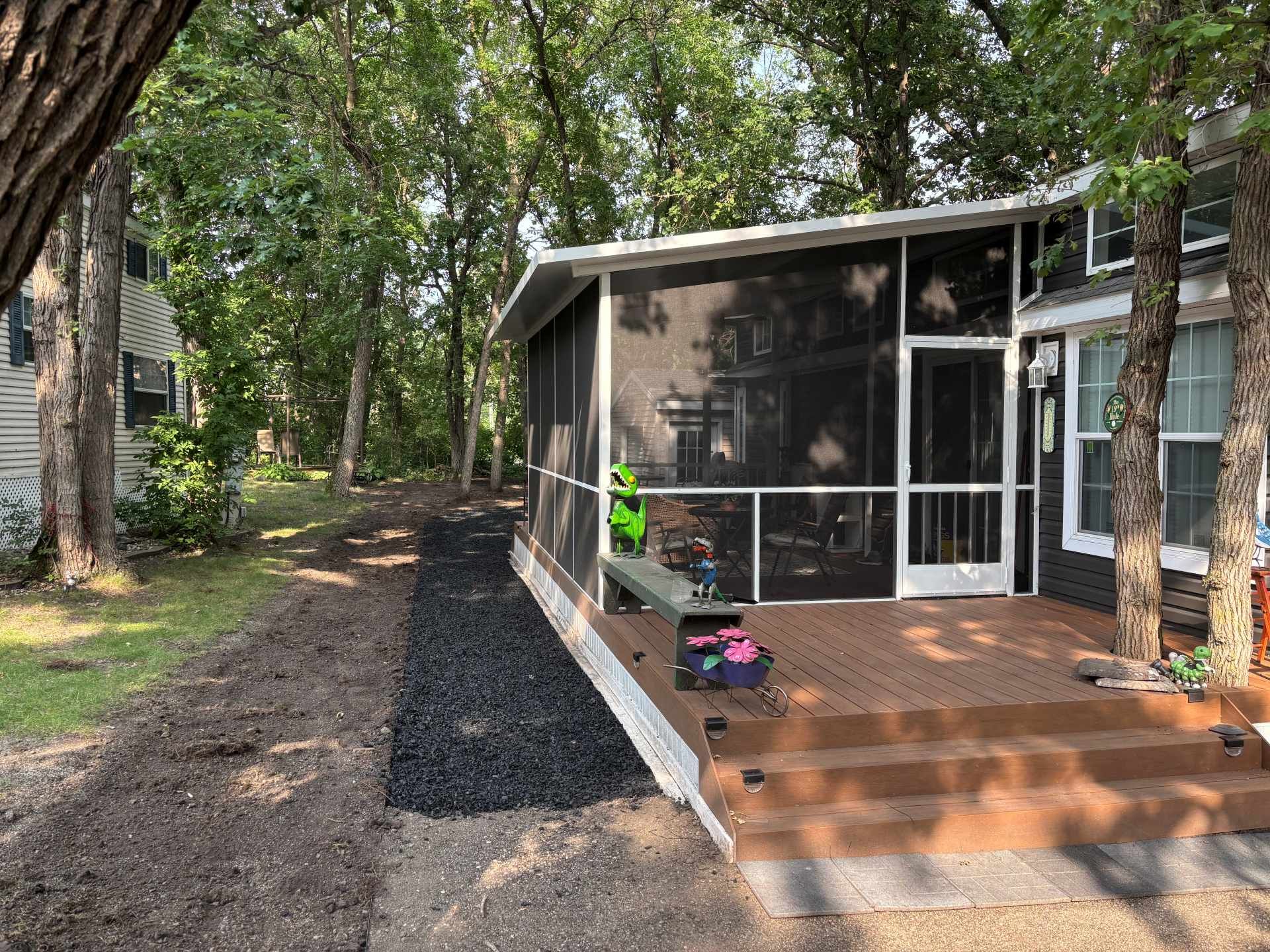 A cabin with a screened-in porch and wooden deck sits in a wooded yard with a gravel pathway along the side.