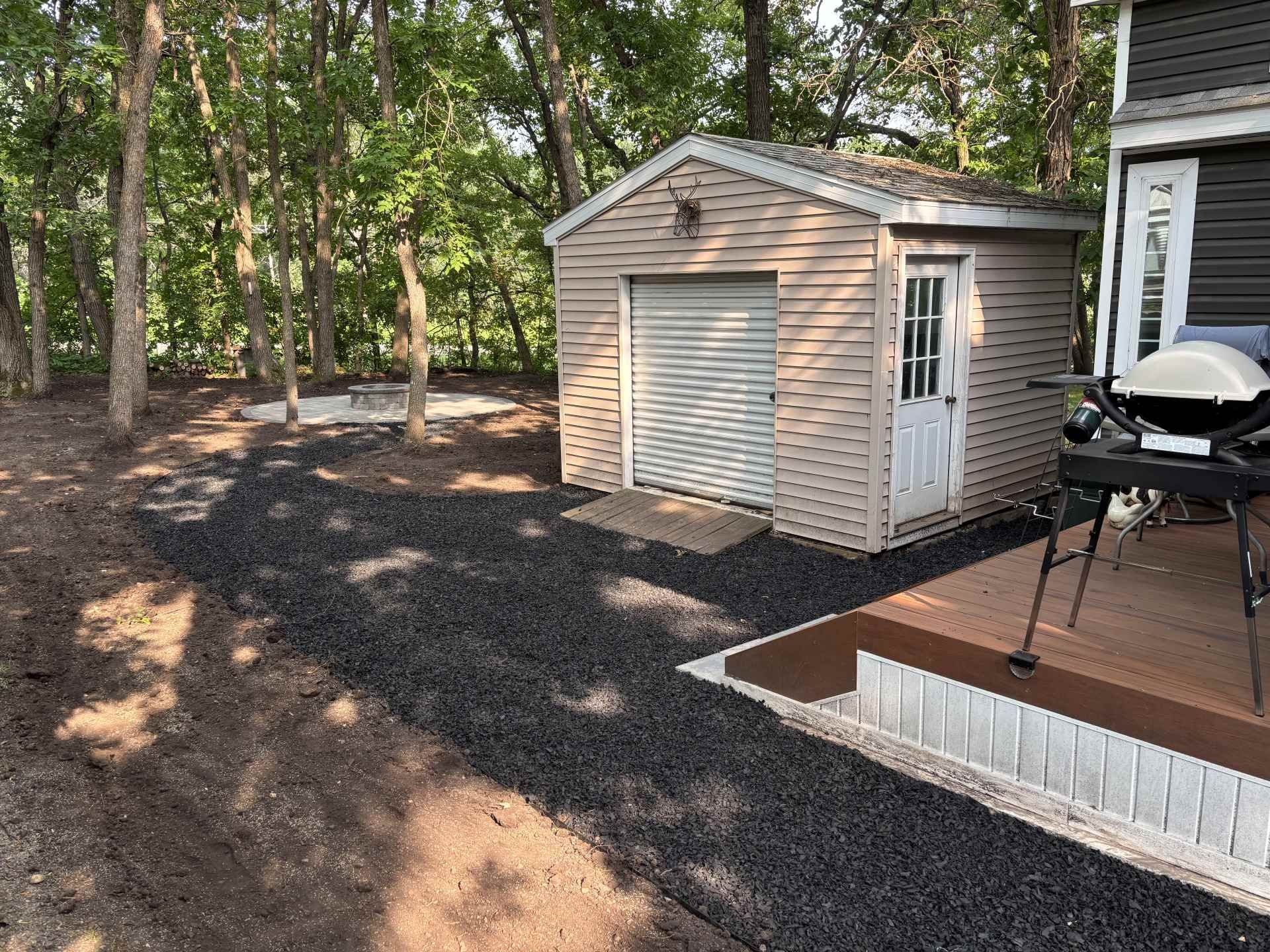 A beige shed sits in a wooded backyard next to a raised deck with a grill and a dark gravel pathway leading to a fire pit.