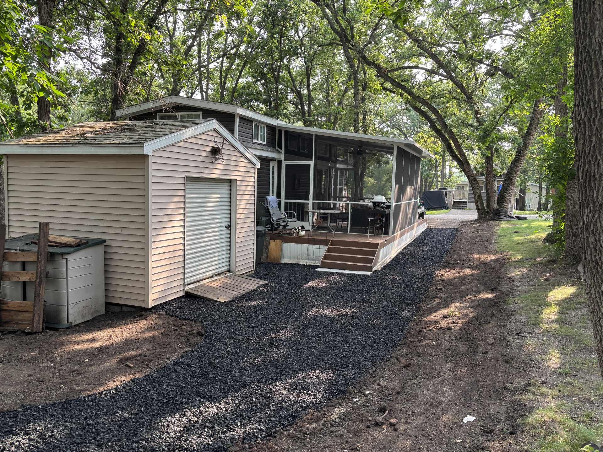 A tan storage shed and a mobile home with a screened-in porch, set on a gravel driveway surrounded by trees.