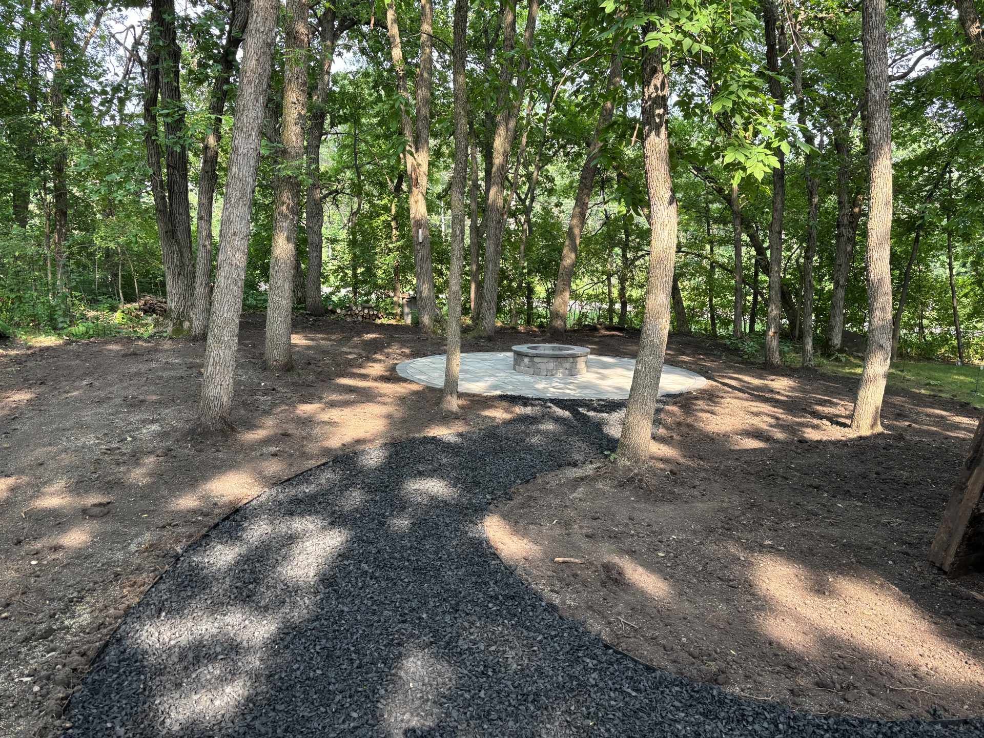 A fire pit area in a wooded clearing with a dark gravel path leading to a light-colored circular gravel patio.