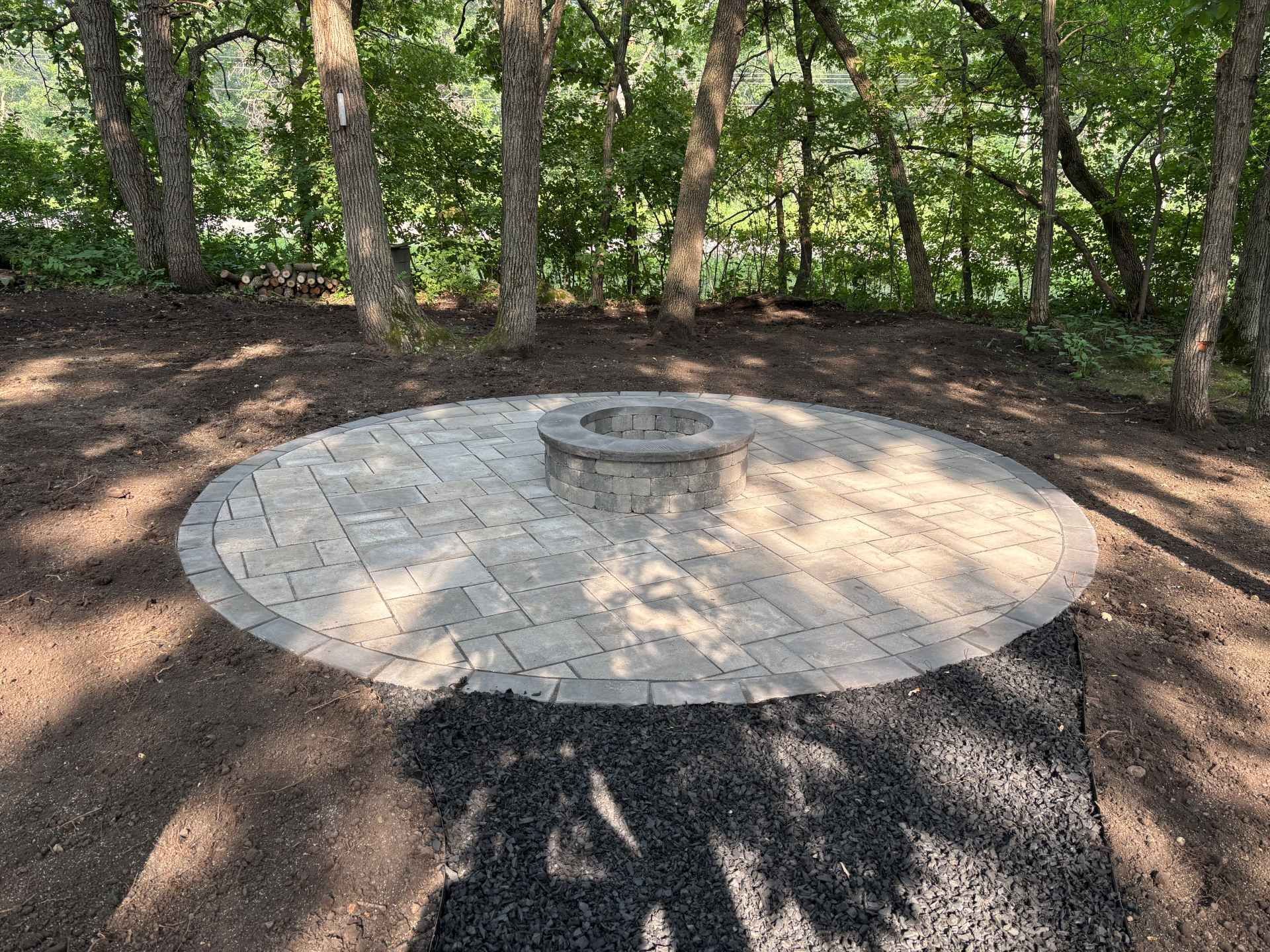A round, stone-paved patio with a central circular fire pit, set in a wooded area with patches of dirt and gravel.