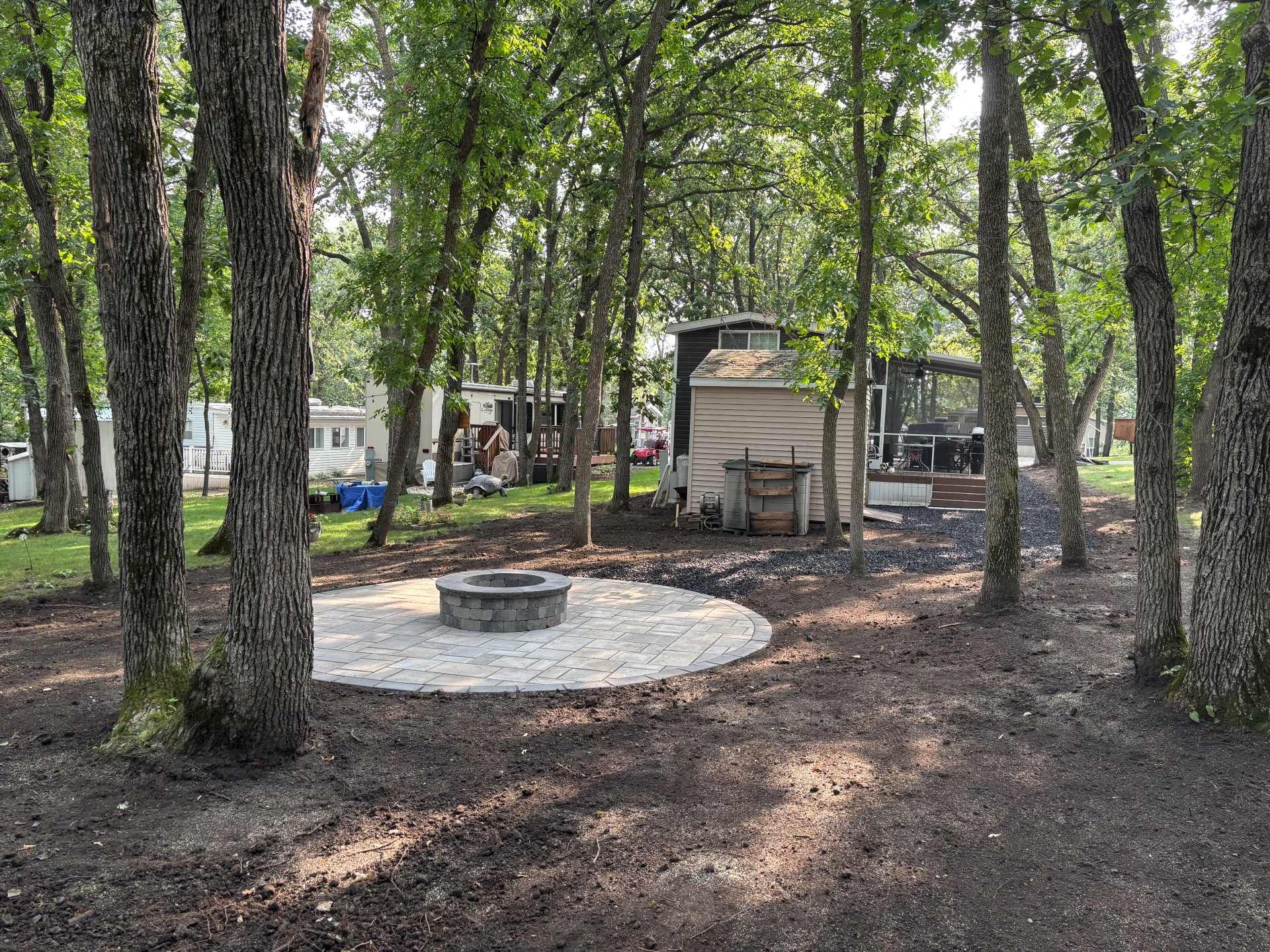 A stone fire pit centered on a paved patio in a wooded yard with nearby mobile homes and trees.