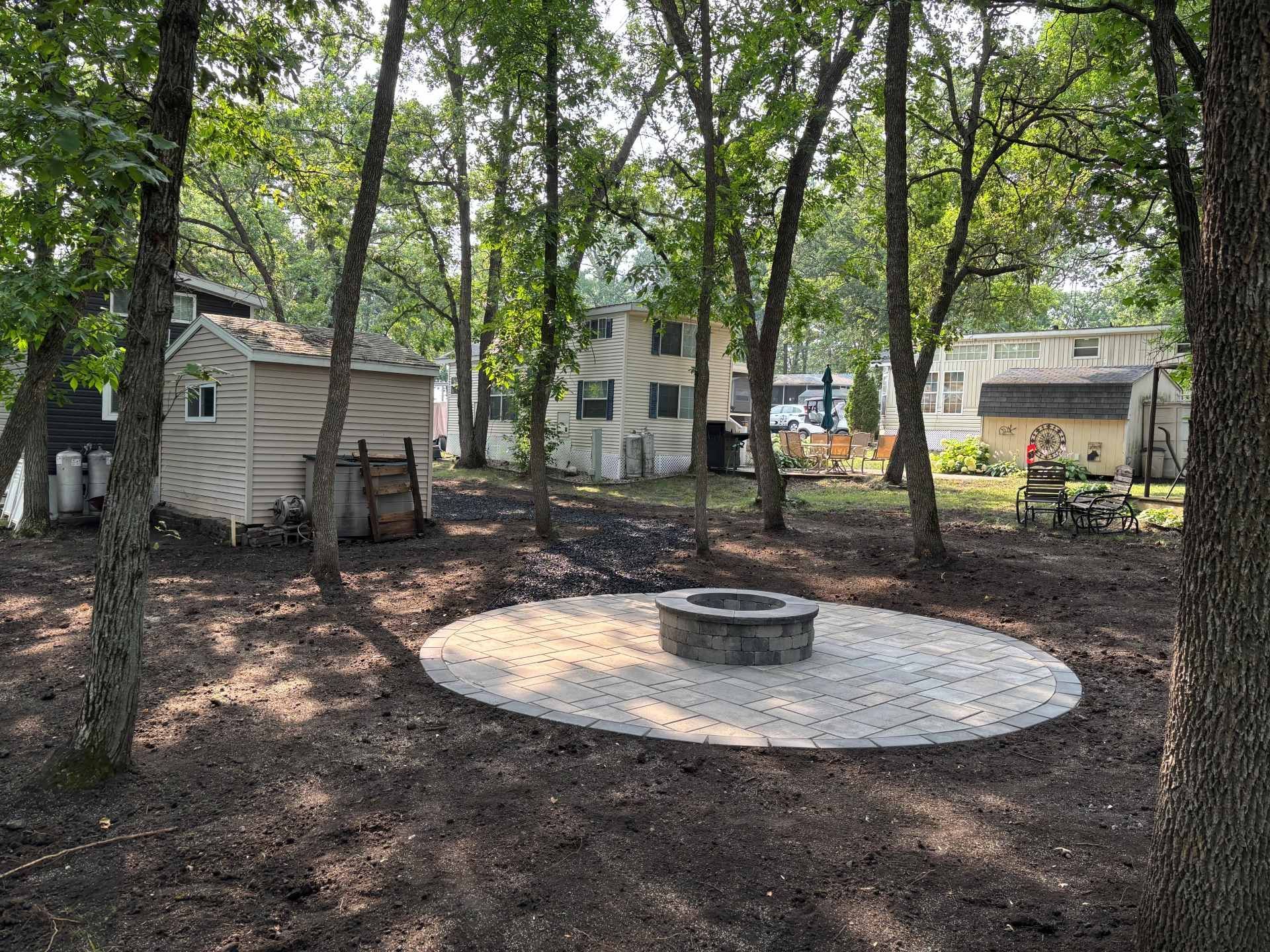 A stone fire pit sits in the center of a circular paver patio within a wooded backyard, flanked by two small outbuildings.