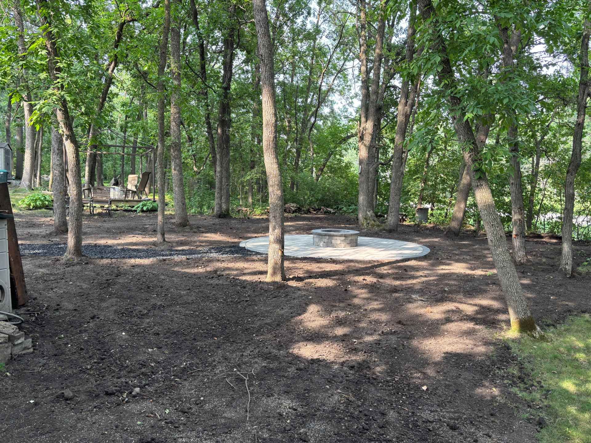 A circular gravel fire pit area sits in a wooded backyard with dark soil and trees.