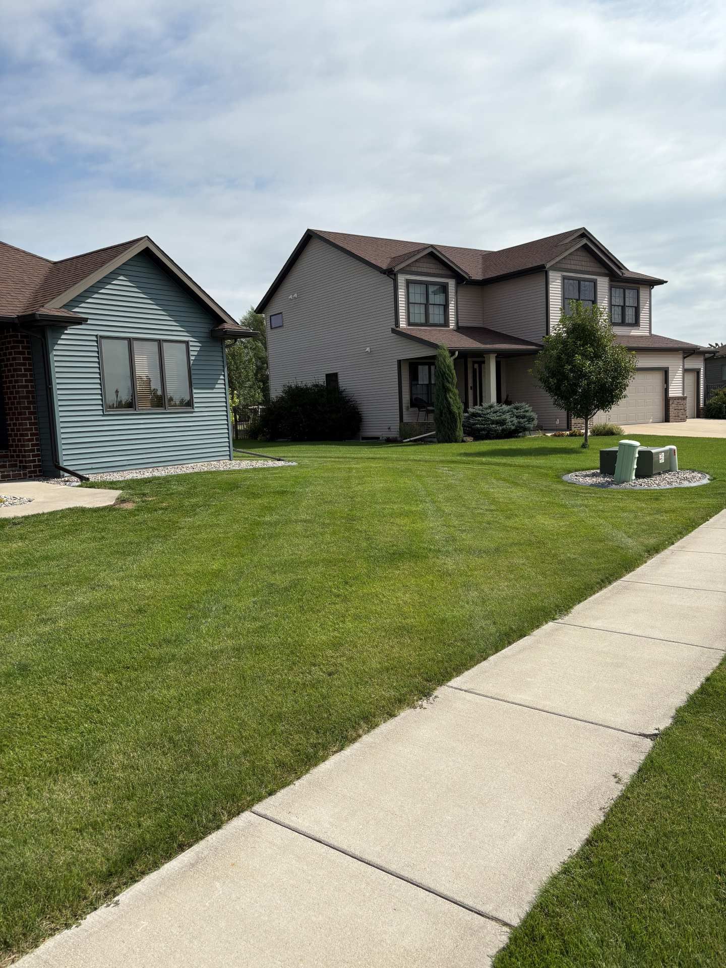 A light gray, two-story house with a blue-gray siding structure in the foreground, separated by a green lawn and sidewalk.