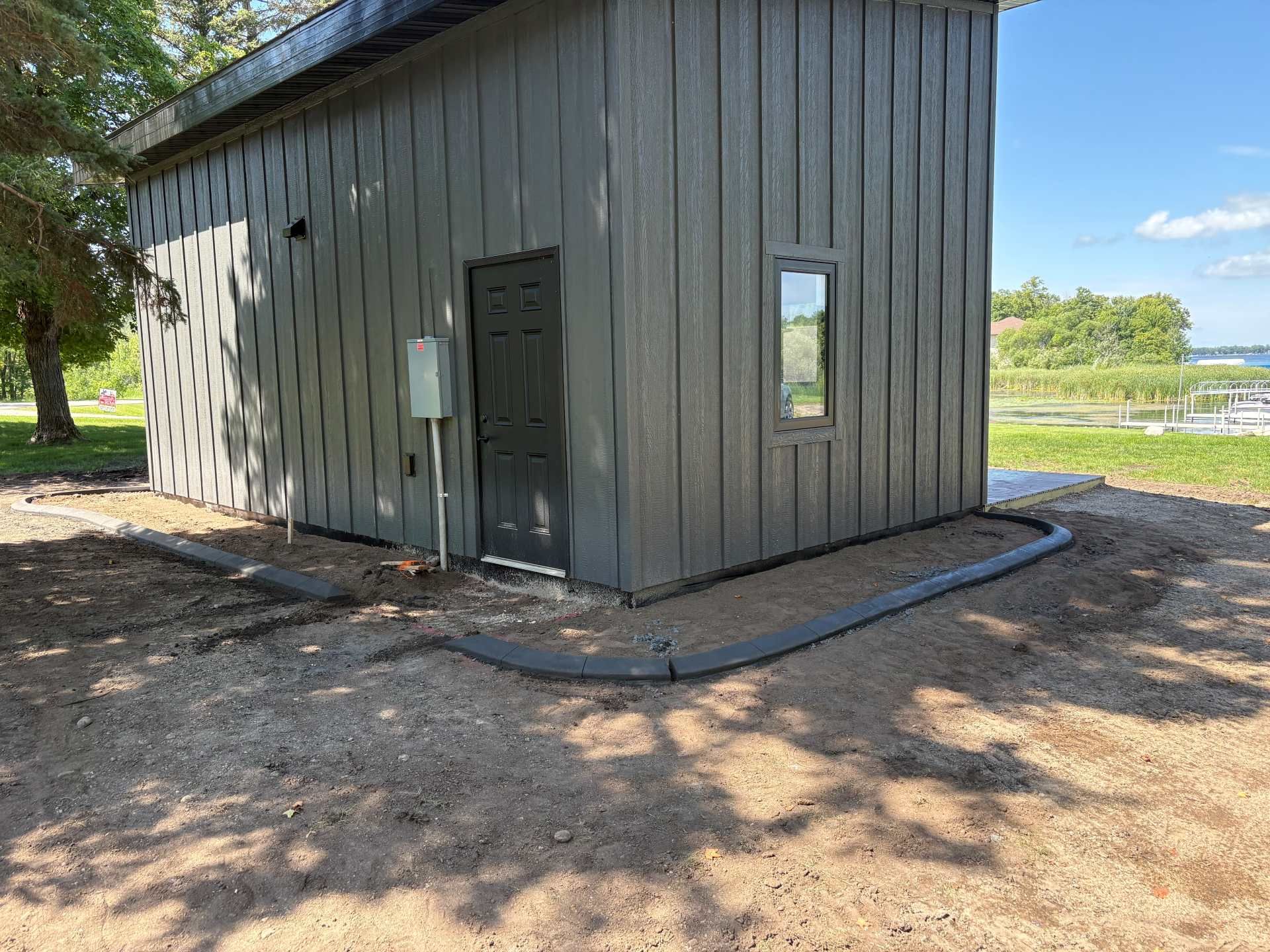 A dark gray modern shed with vertical siding, a door, and a single window, set on a dirt lot with black garden edging.