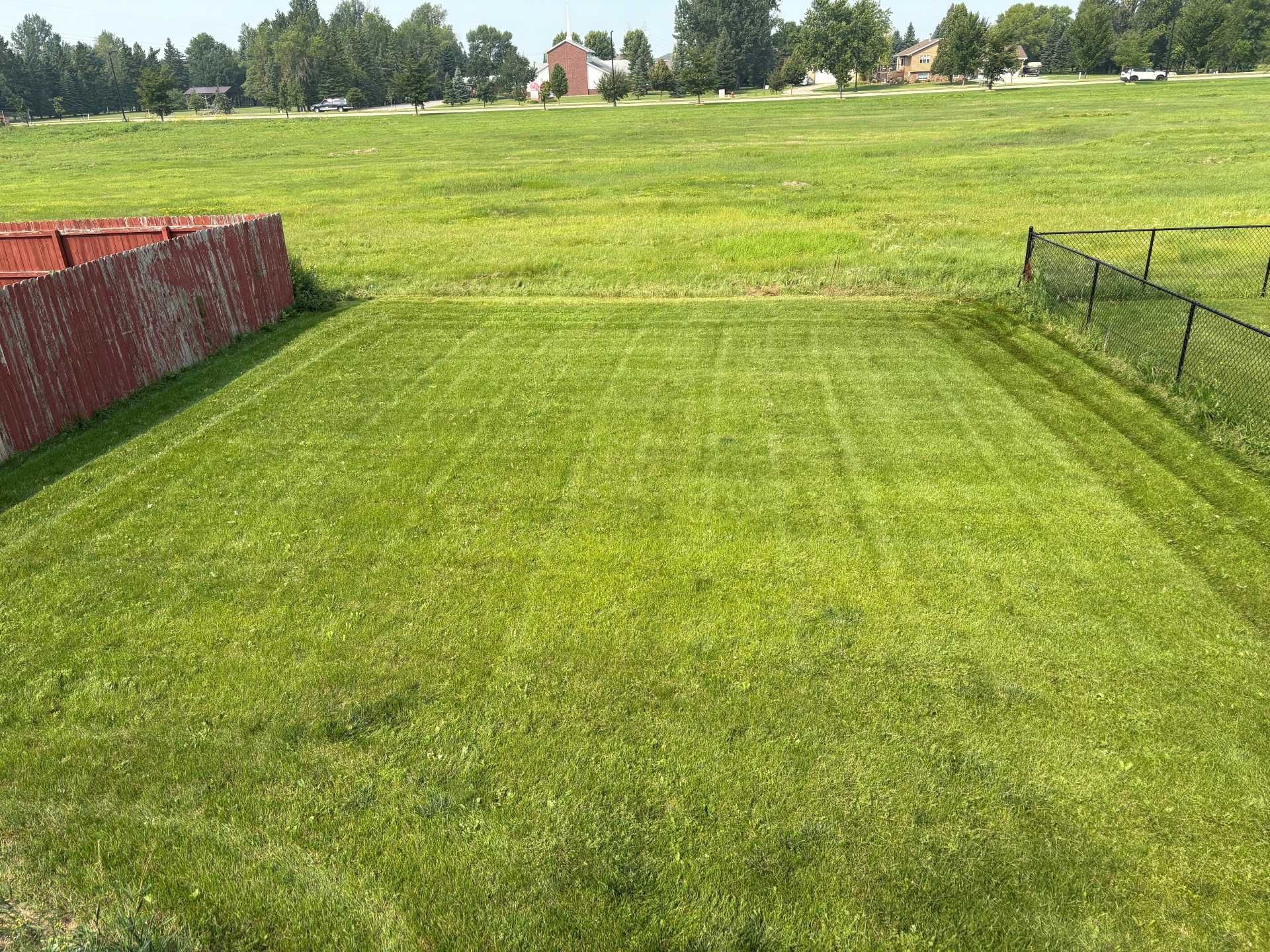 A recently mowed rectangular lawn bordered by a red fence on the left and a chain-link fence on the right.