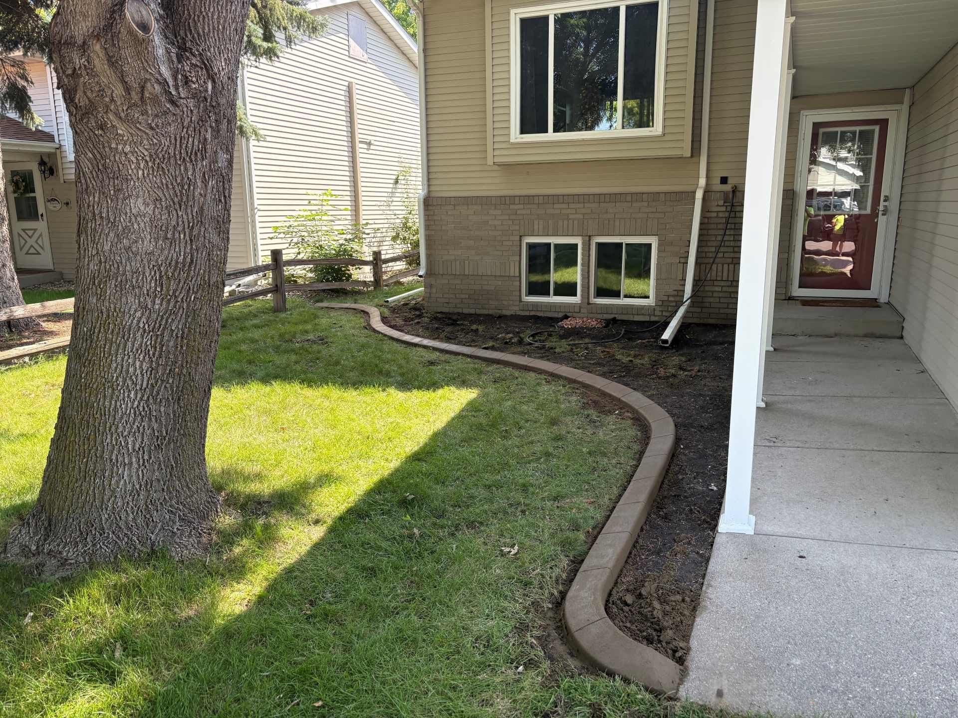 A concrete curb separates a lawn from a garden bed along the side of a tan house with a red front door.