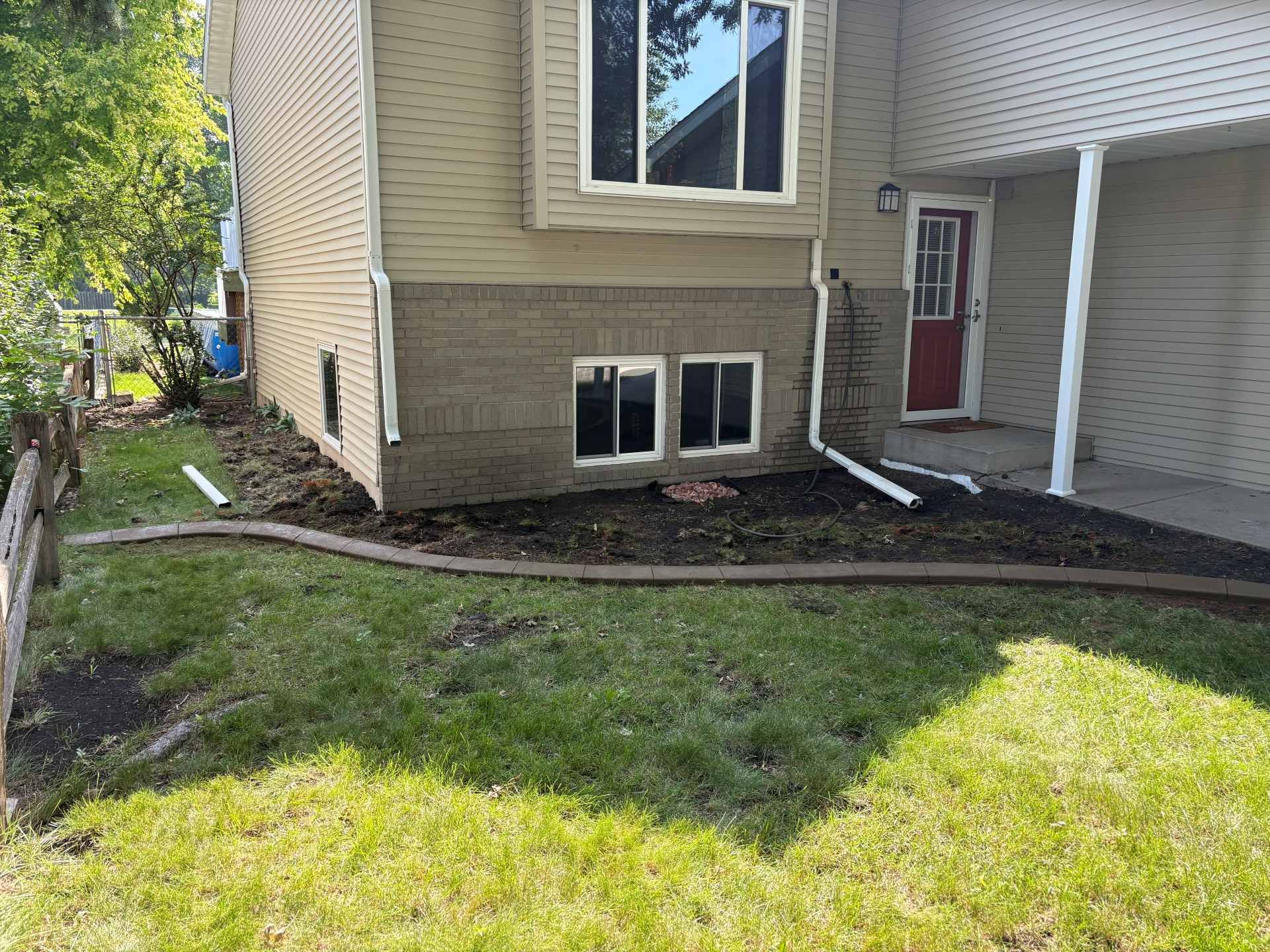 A beige house with a brick foundation, a white-trimmed bay window, and a mulched garden bed in the front yard.