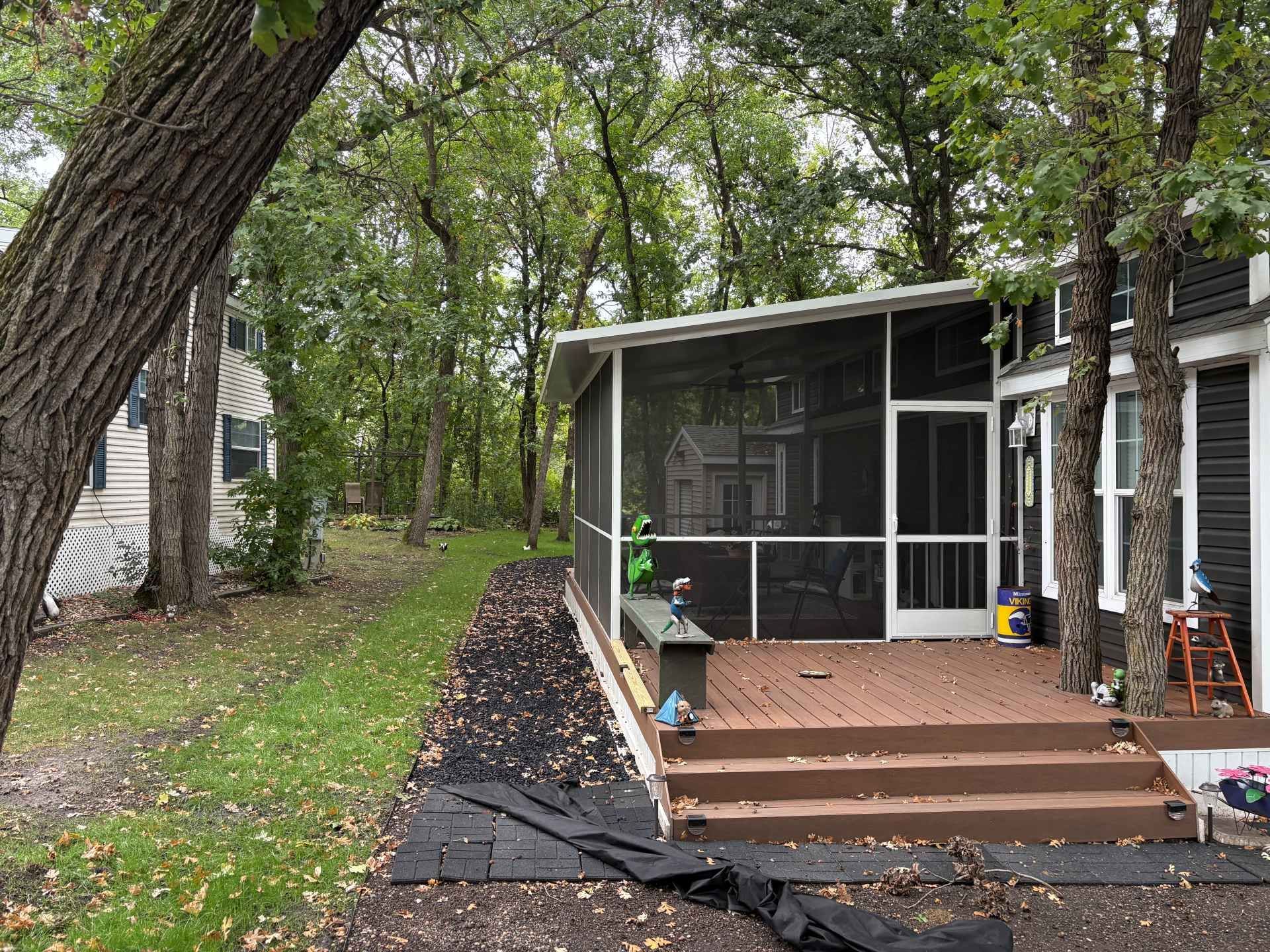 A wooden deck with a screened-in porch sits beside a dark house and trees, with a yard covered in fallen autumn leaves.