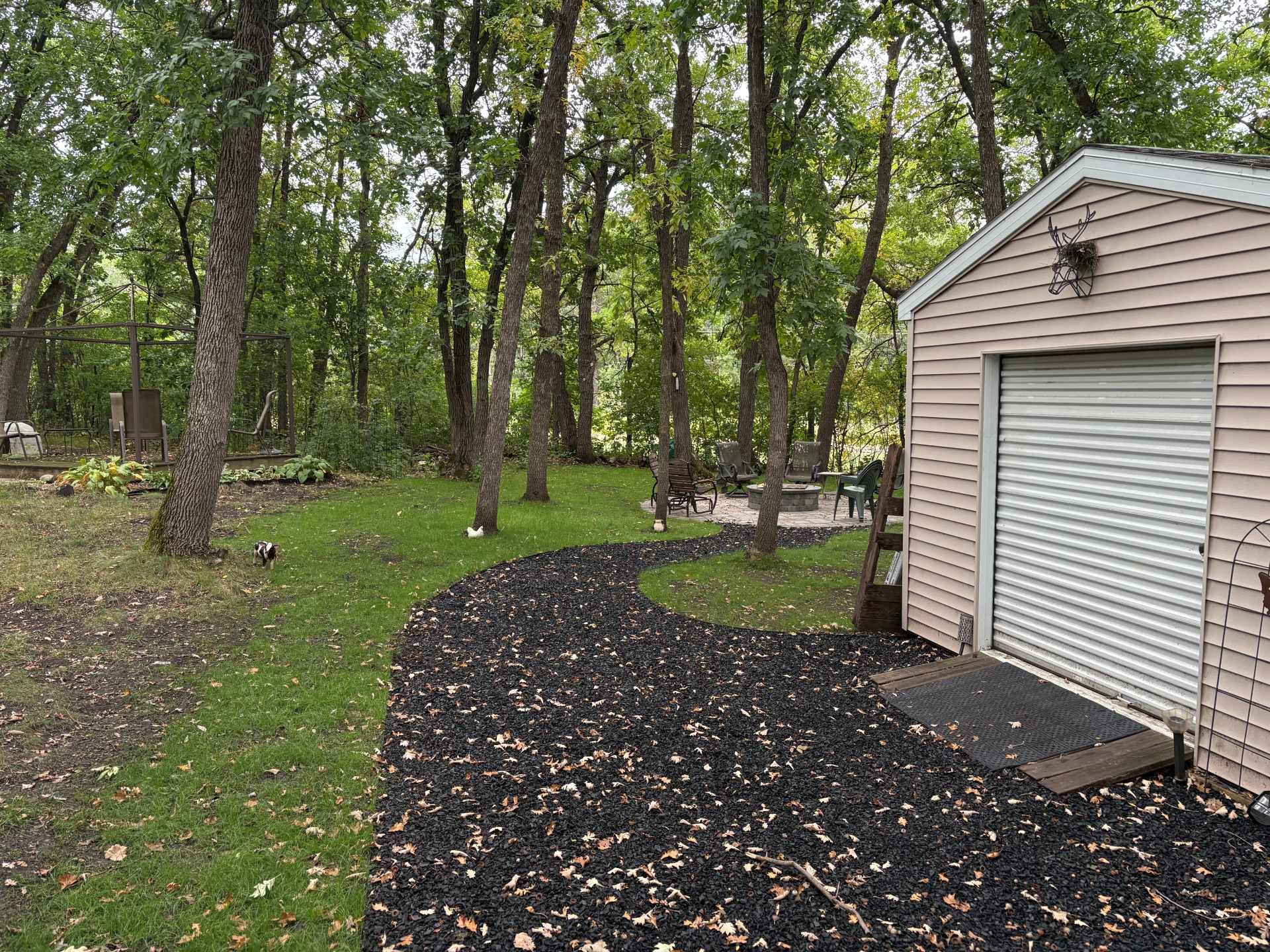 A black mulch path leads through a grassy, wooded backyard beside a light-colored storage shed.