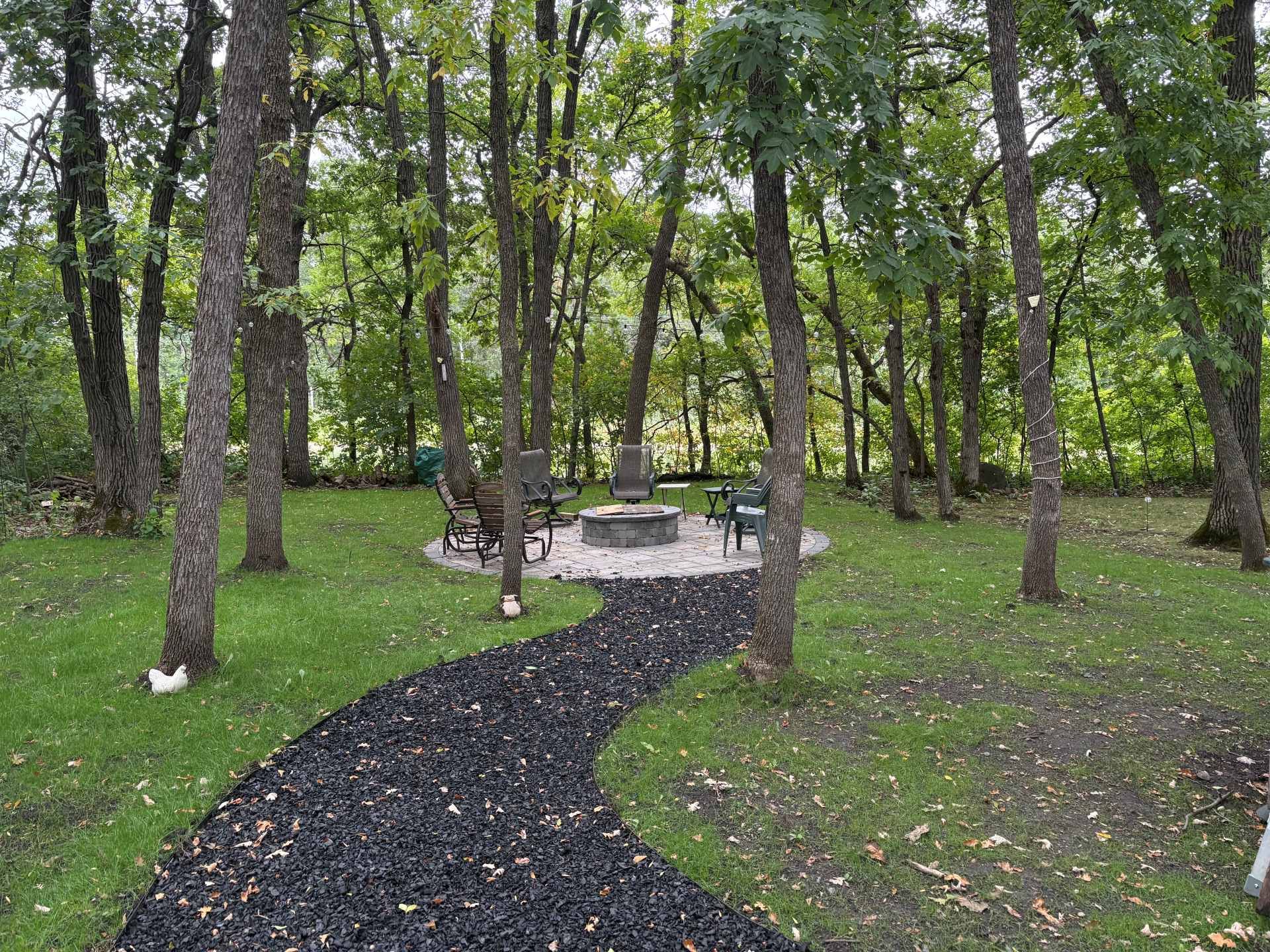 A curving black gravel path leads to a circular stone fire pit with chairs in a lush, wooded backyard.
