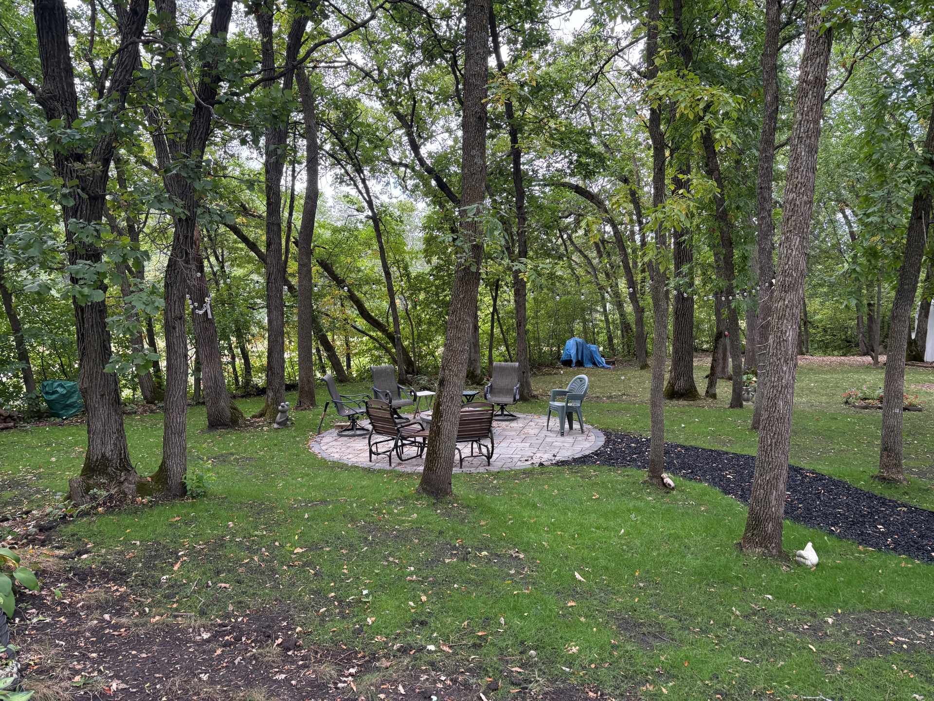 A stone circular fire pit area with outdoor chairs sits in a grassy, wooded backyard with a dark gravel path.