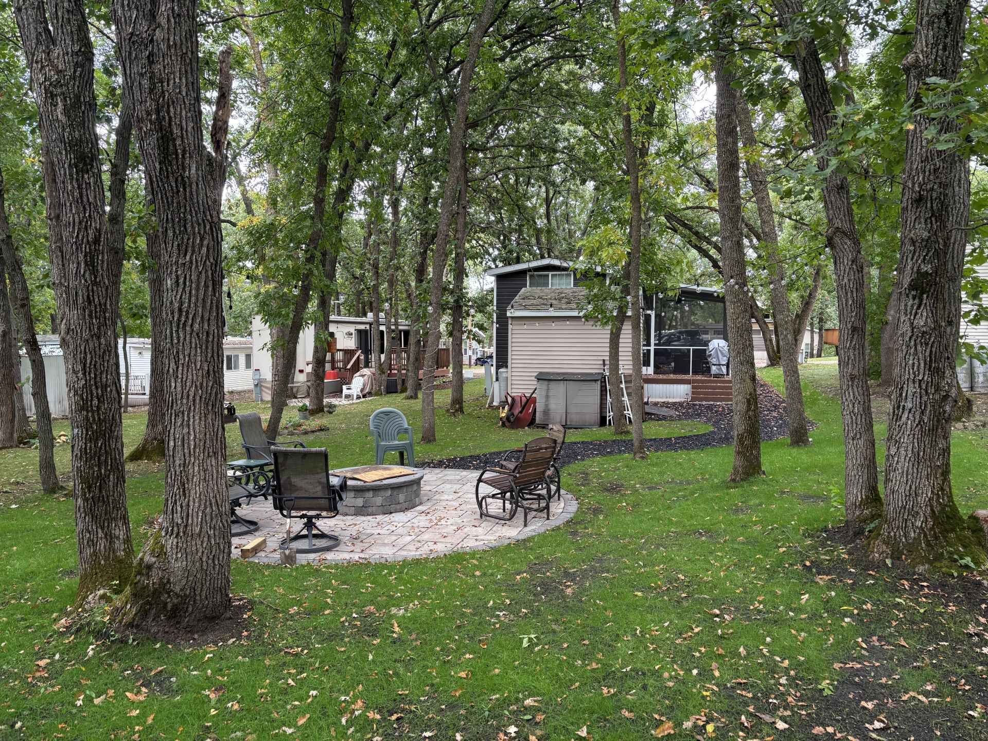 A backyard fire pit area with stone patio, chairs, and trees, featuring a mobile home in the background.