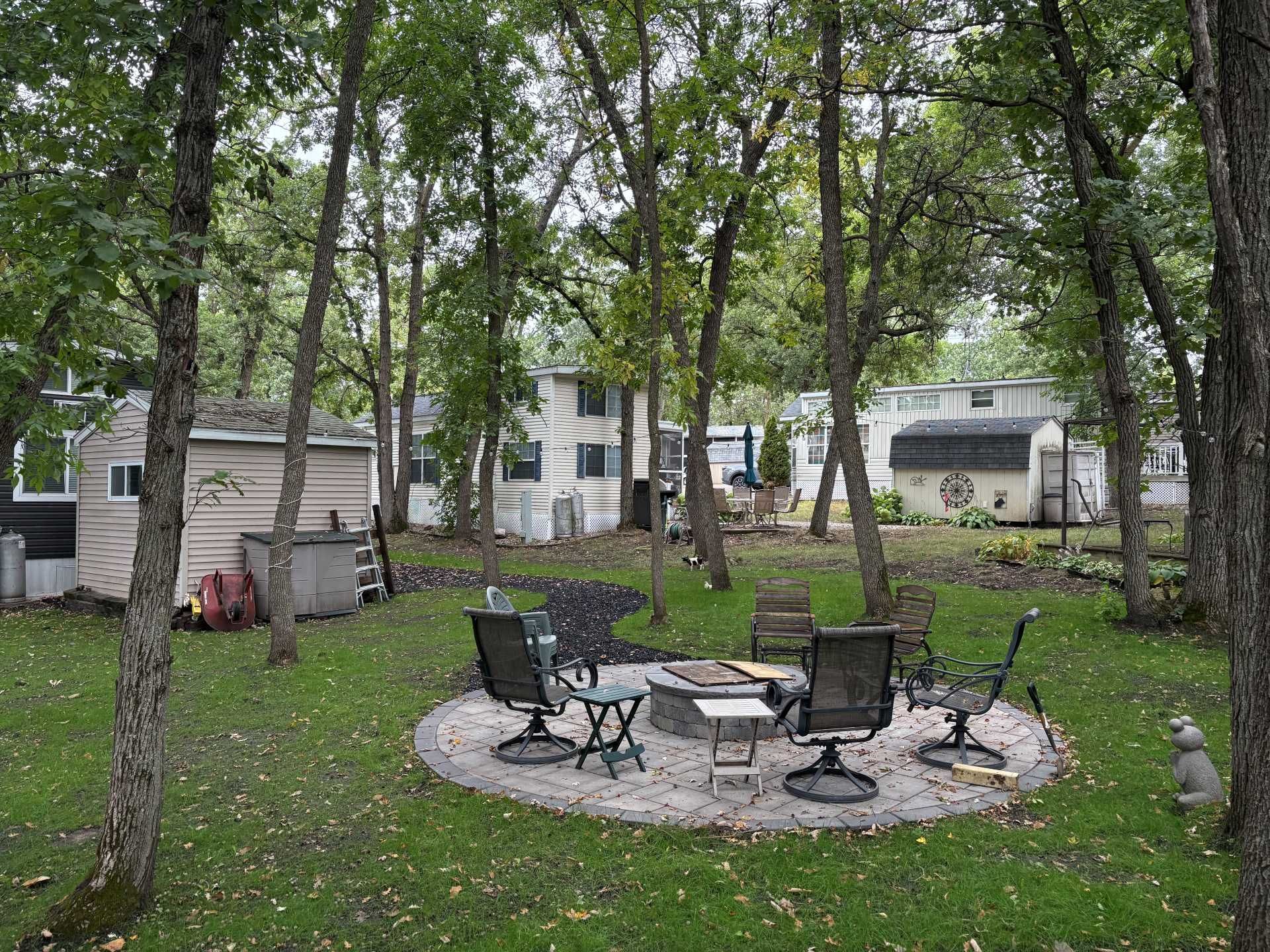 A stone fire pit surrounded by outdoor chairs in a grassy, tree-filled backyard with small structures in the background.