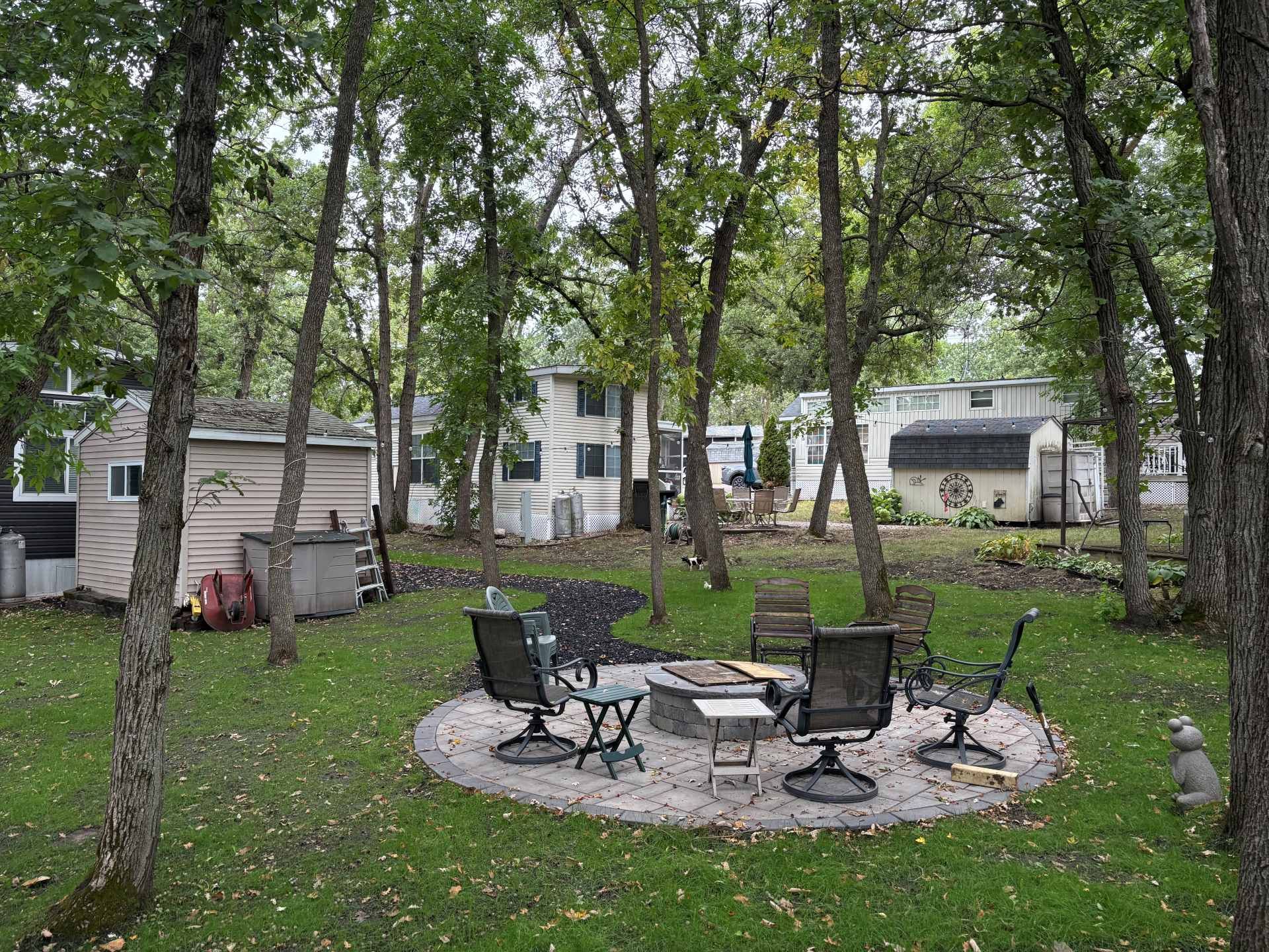 A circular stone fire pit with outdoor chairs in a grassy backyard surrounded by trees, with small sheds in the background.