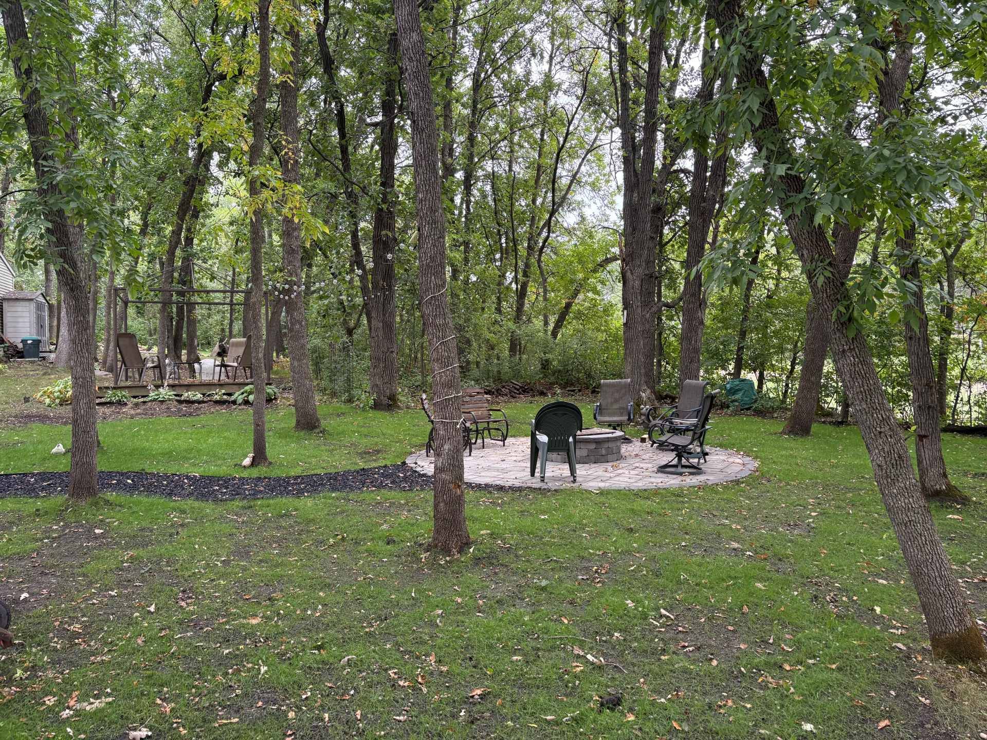 A fire pit with surrounding black chairs on a circular stone patio in a grassy, wooded backyard.