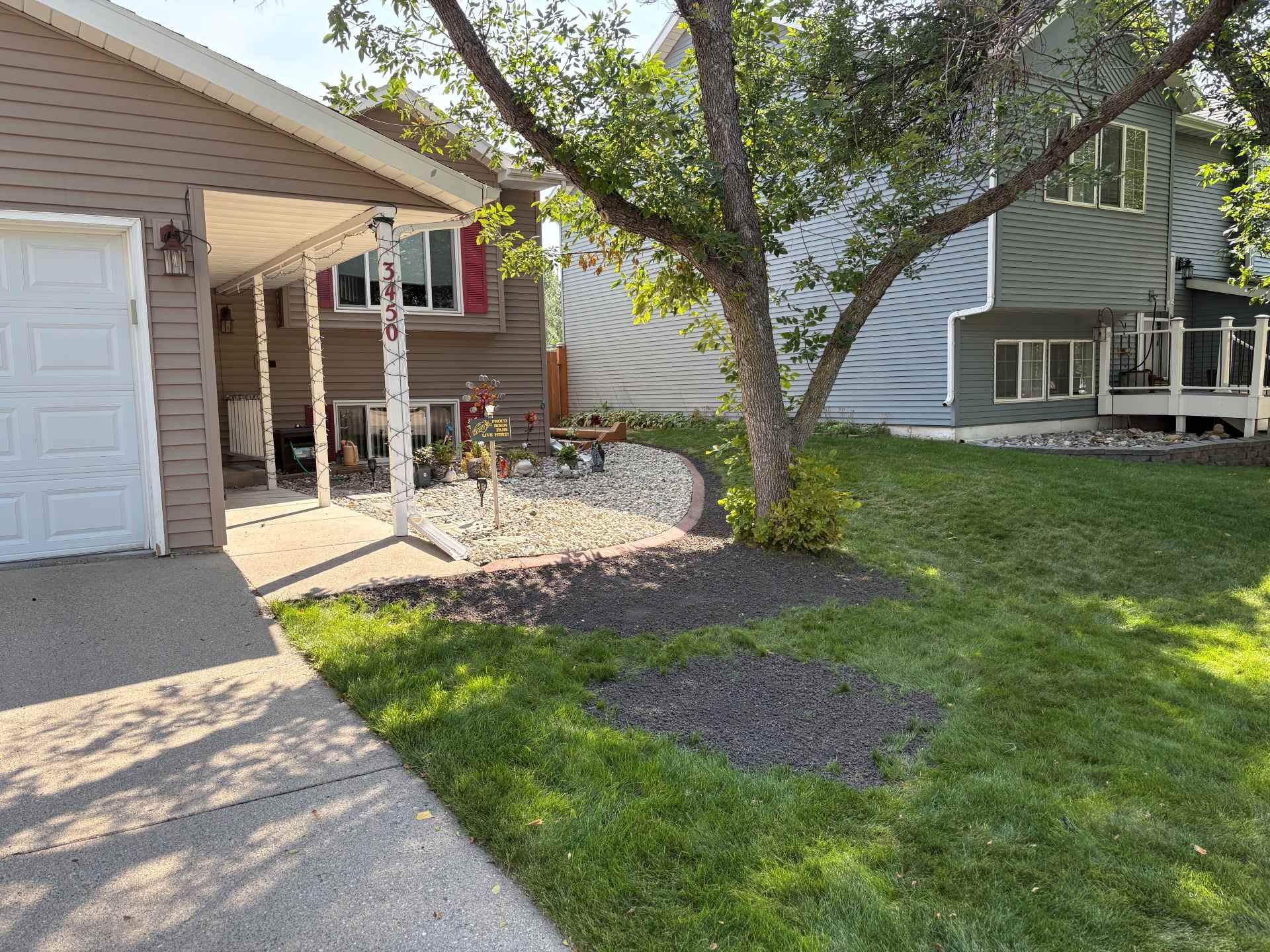 A residential exterior view showing a driveway, a brown home with a covered porch, a large tree, and a grassy front lawn.