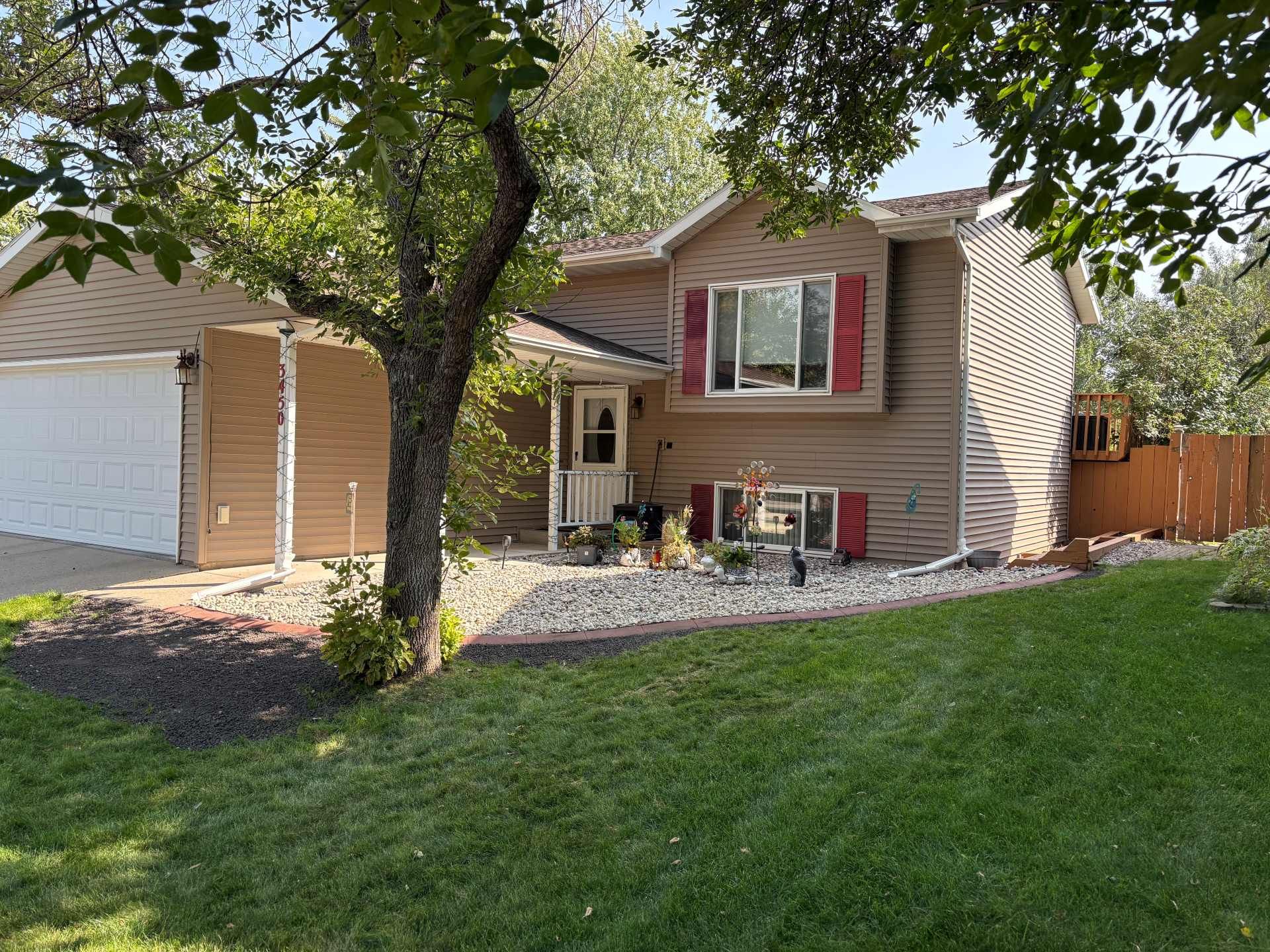 A multi-level suburban home with tan vinyl siding, white garage doors, and red window shutters, seen from a lawn.