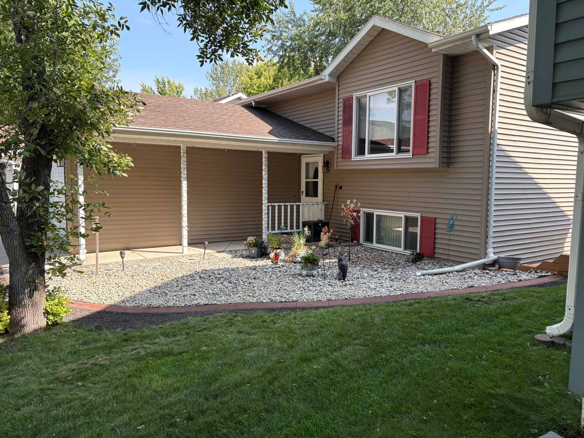 A tan split-level house with red shutters, a gravel front yard, a covered porch, and a patch of green grass in front.