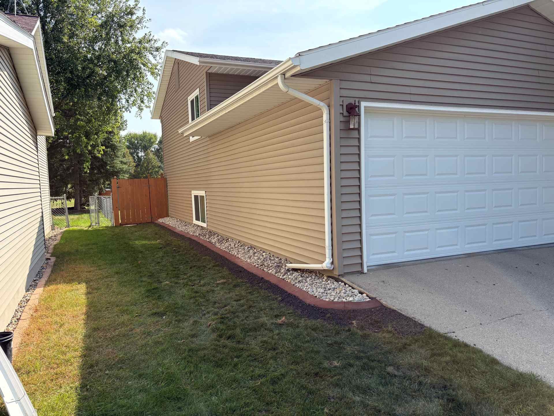 Side view of a tan-sided house with a white garage door, gravel landscaping, and a fenced backyard on a sunny day.