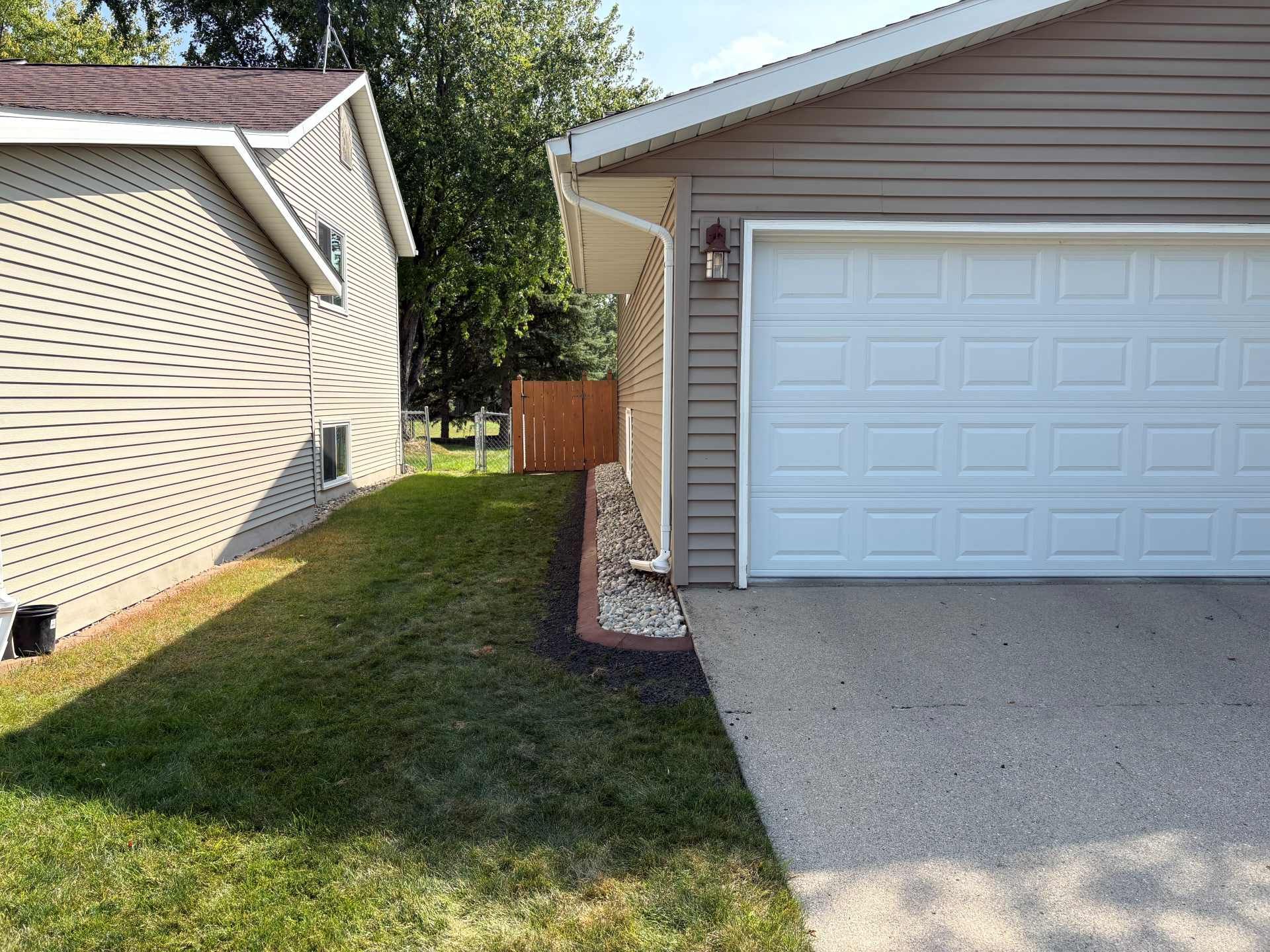 A gravel garden bed runs along the side of a beige house with a garage, adjacent to a grassy yard and a neighboring house.