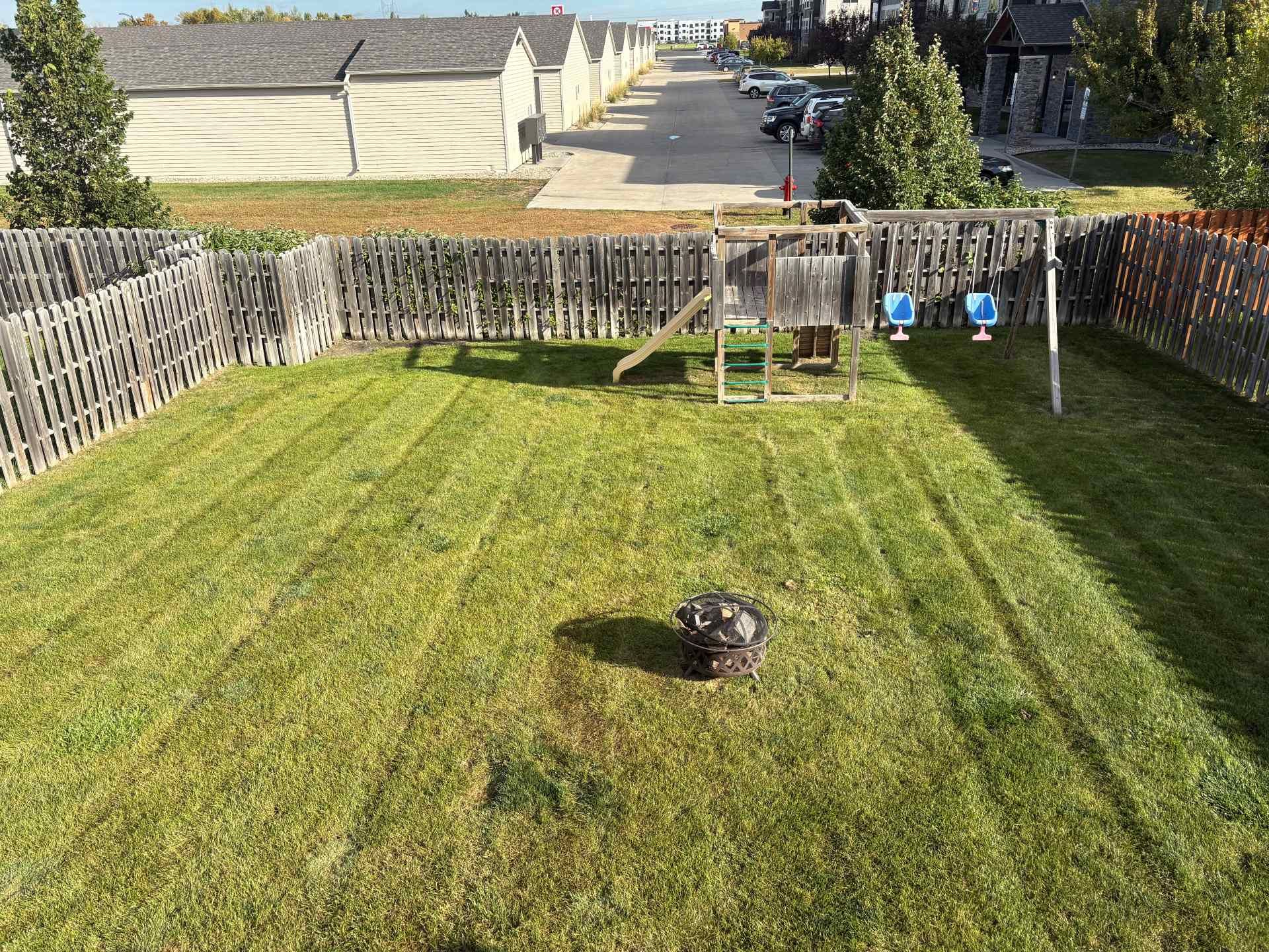 A grassy backyard with a play set, swings, a fire pit, and a wooden fence, viewed from an elevated perspective.