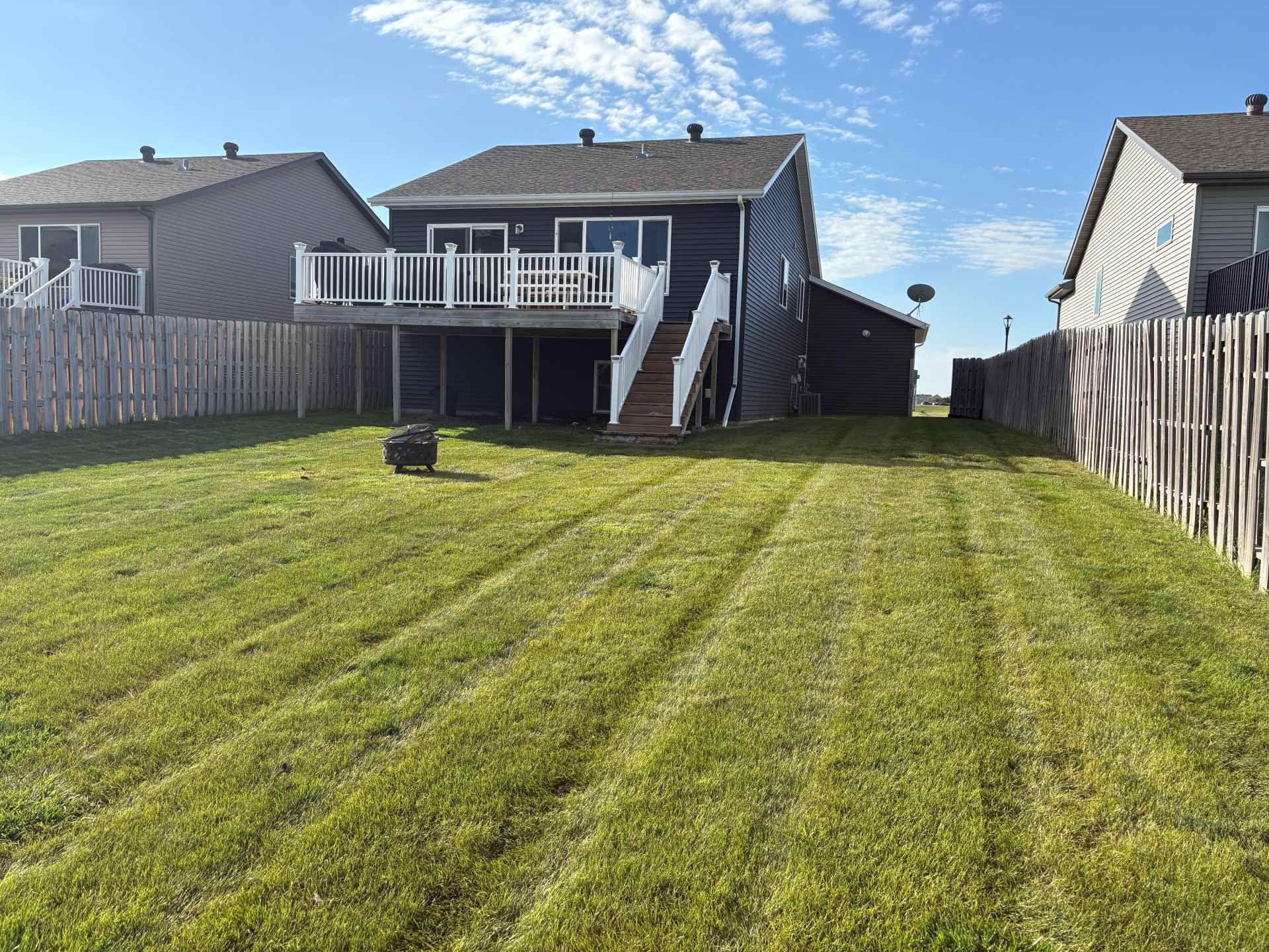 A backyard with freshly mowed green grass, wooden fences, and a multi-level house with a white deck and stairs.