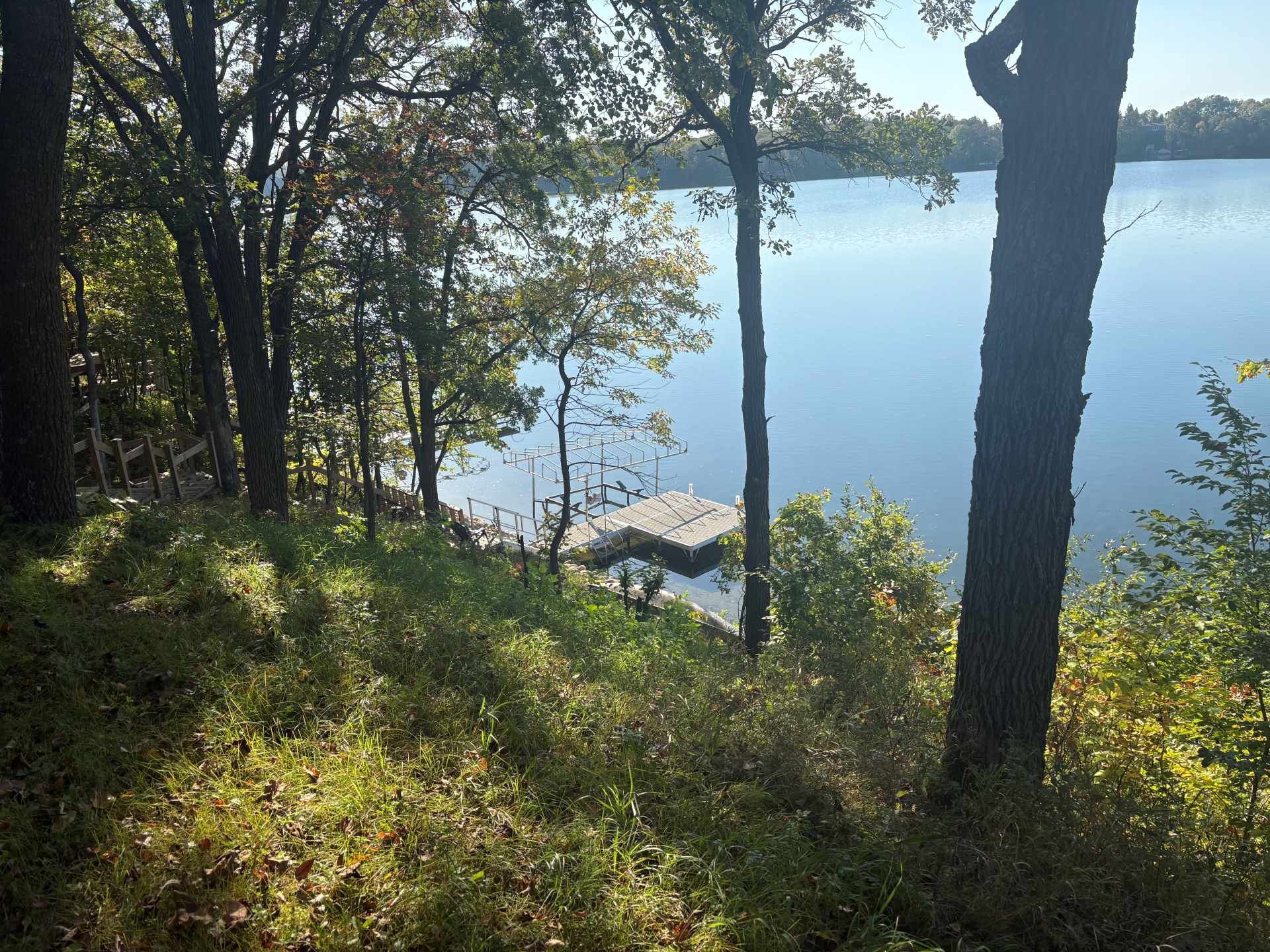 A view from a grassy, wooded hillside overlooking a calm lake with a wooden boat dock extending into the water.