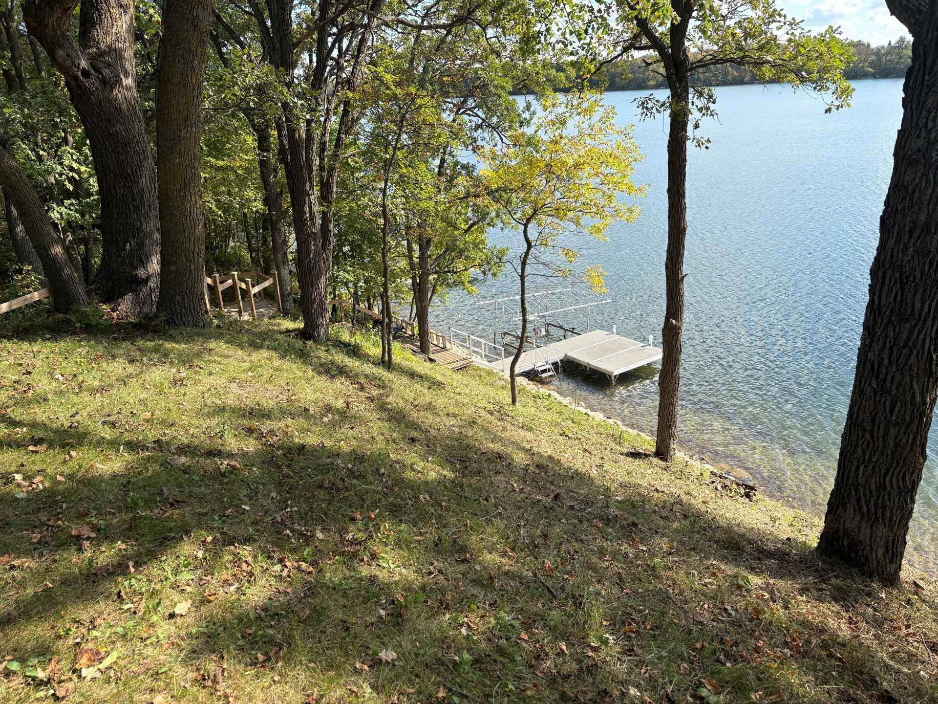 A grassy, tree-lined slope overlooks a calm lake with a small wooden dock extending into the water.