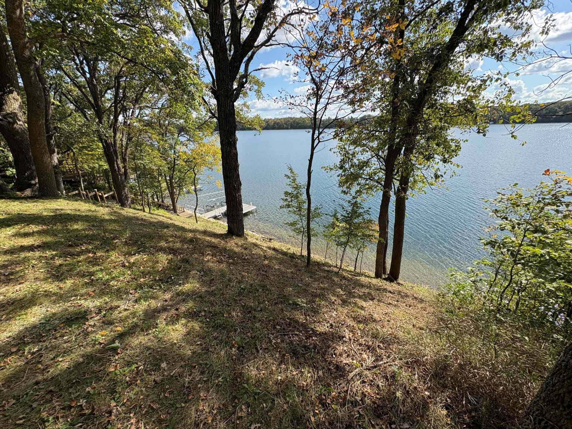 A grassy, sloping lakeshore with trees and foliage in the foreground leading to a calm lake and a small boat dock.
