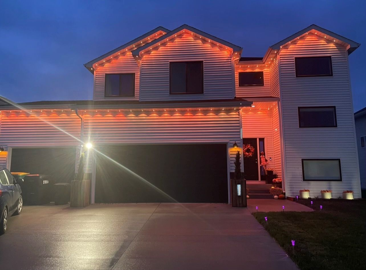 A two-story house at dusk, decorated with glowing orange string lights along the roofline and entryway.