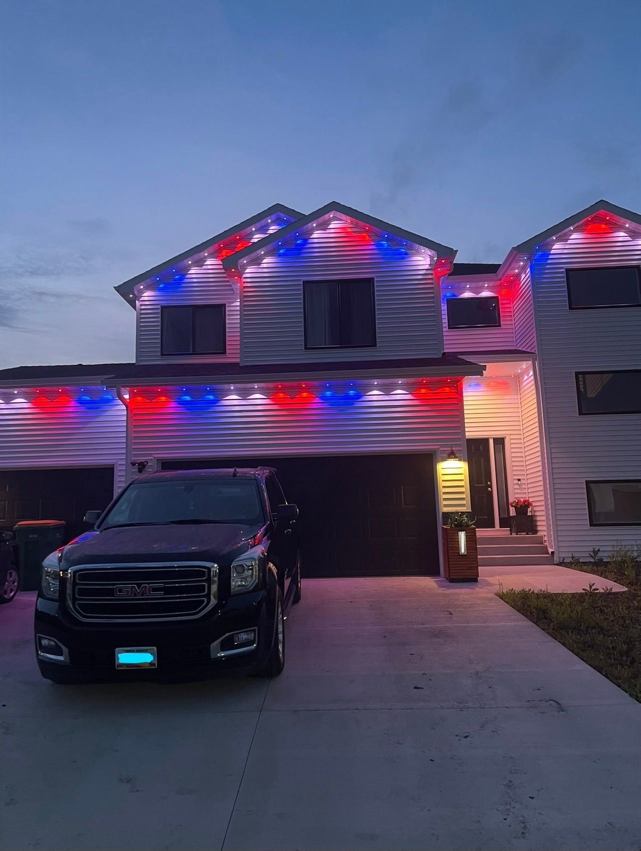 A two-story house at dusk decorated with red, white, and blue exterior lights, with a dark SUV parked in the driveway.