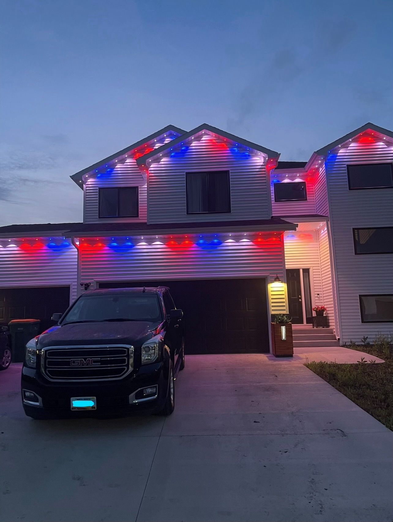 A two-story house at dusk decorated with red, white, and blue lights, with a dark SUV parked in the driveway.